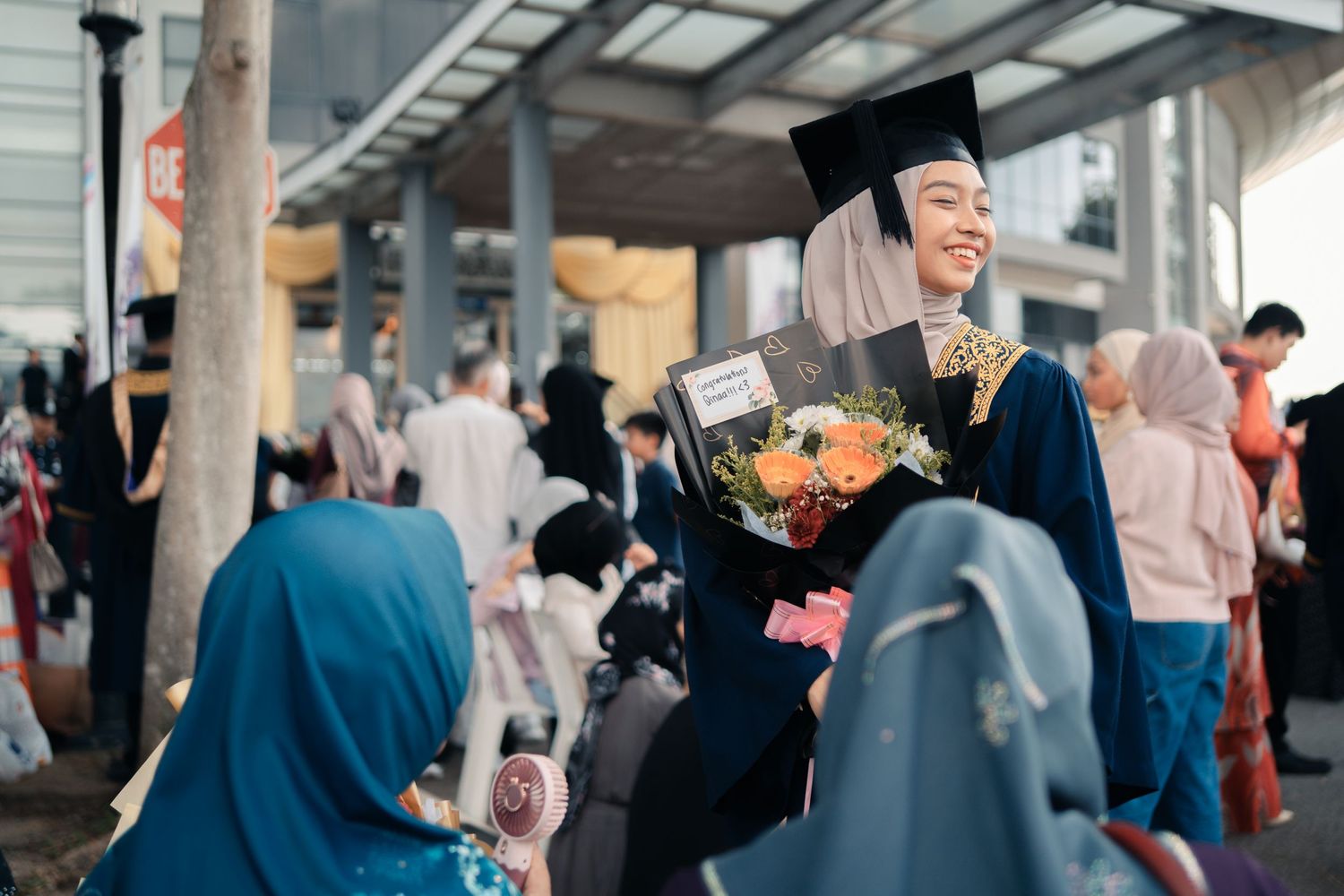 Graduate in blue regalia holding bouquet at outdoor ceremony celebration.