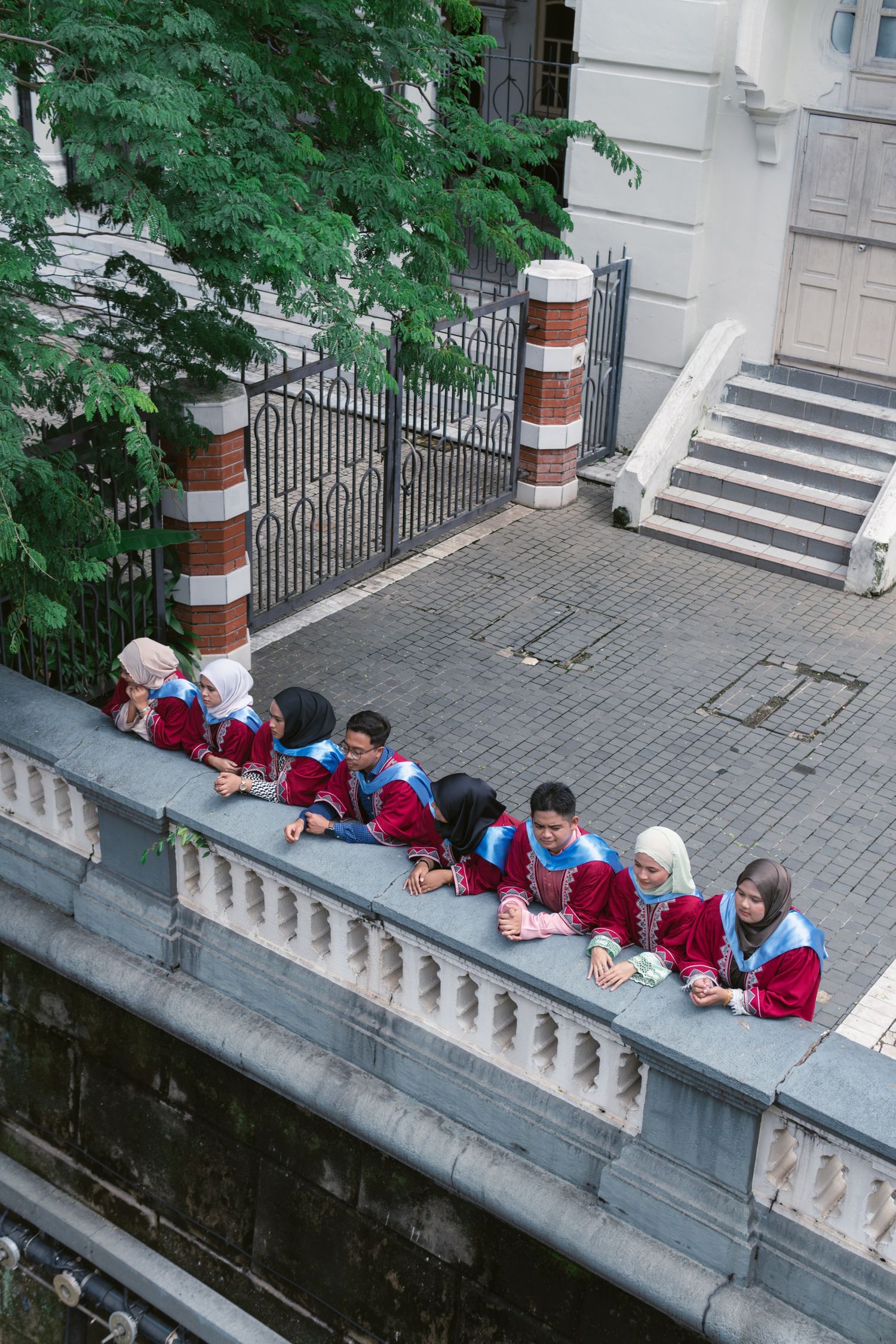 Students in red uniforms sitting in a row on a stone ledge near an ornate gate.