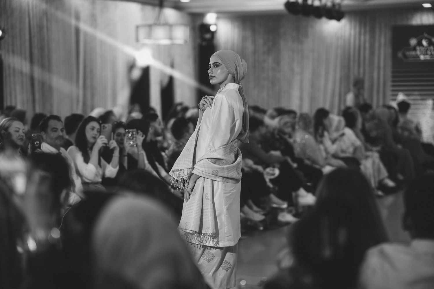 Black and white photo of model walking fashion runway with audience in background.