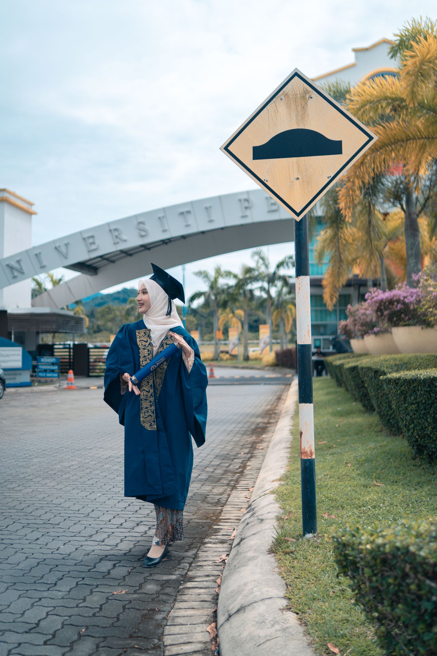 Graduate in blue academic regalia stands next to street sign on university campus.