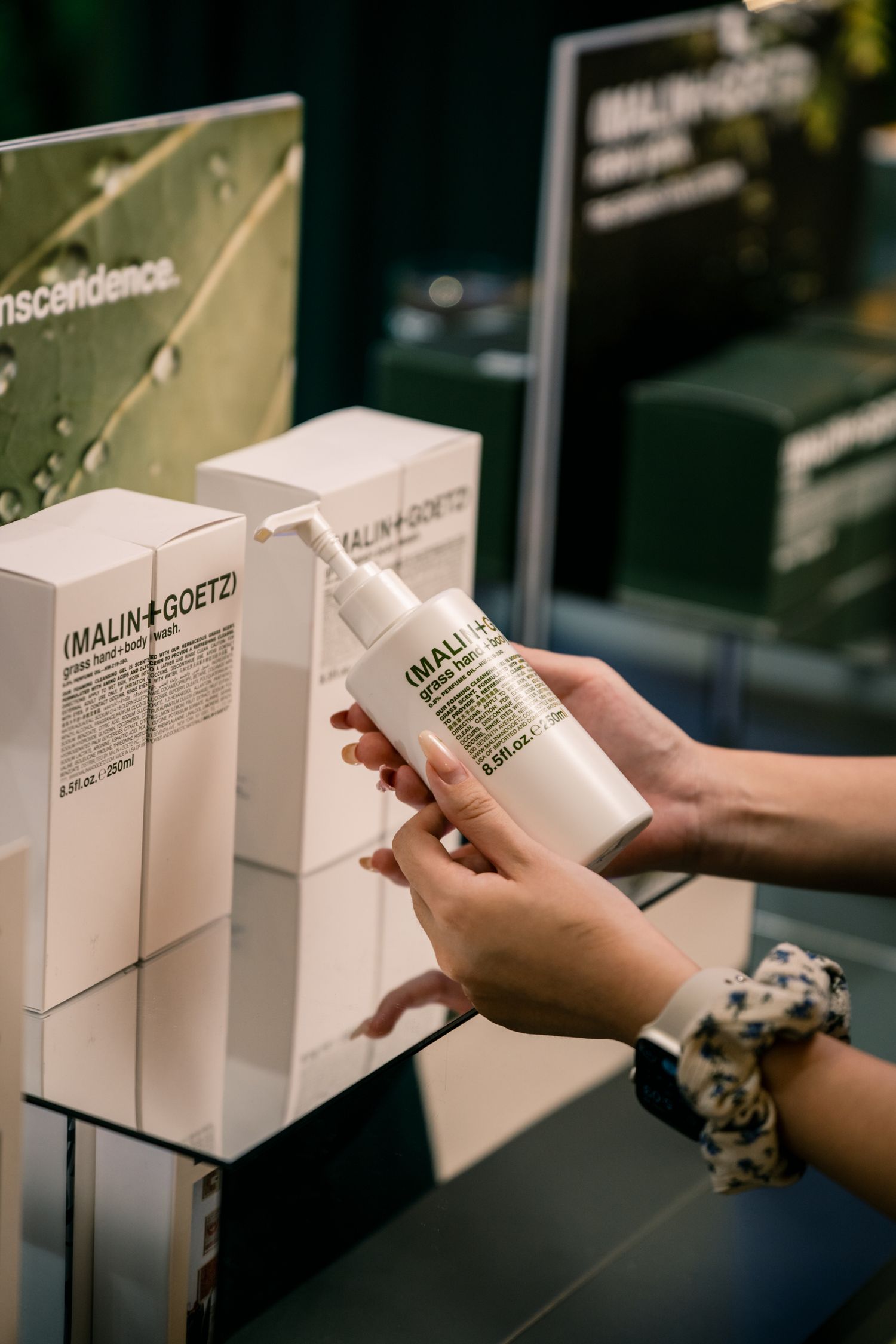 Hands holding Malin+Goetz product bottle against display of white product boxes.