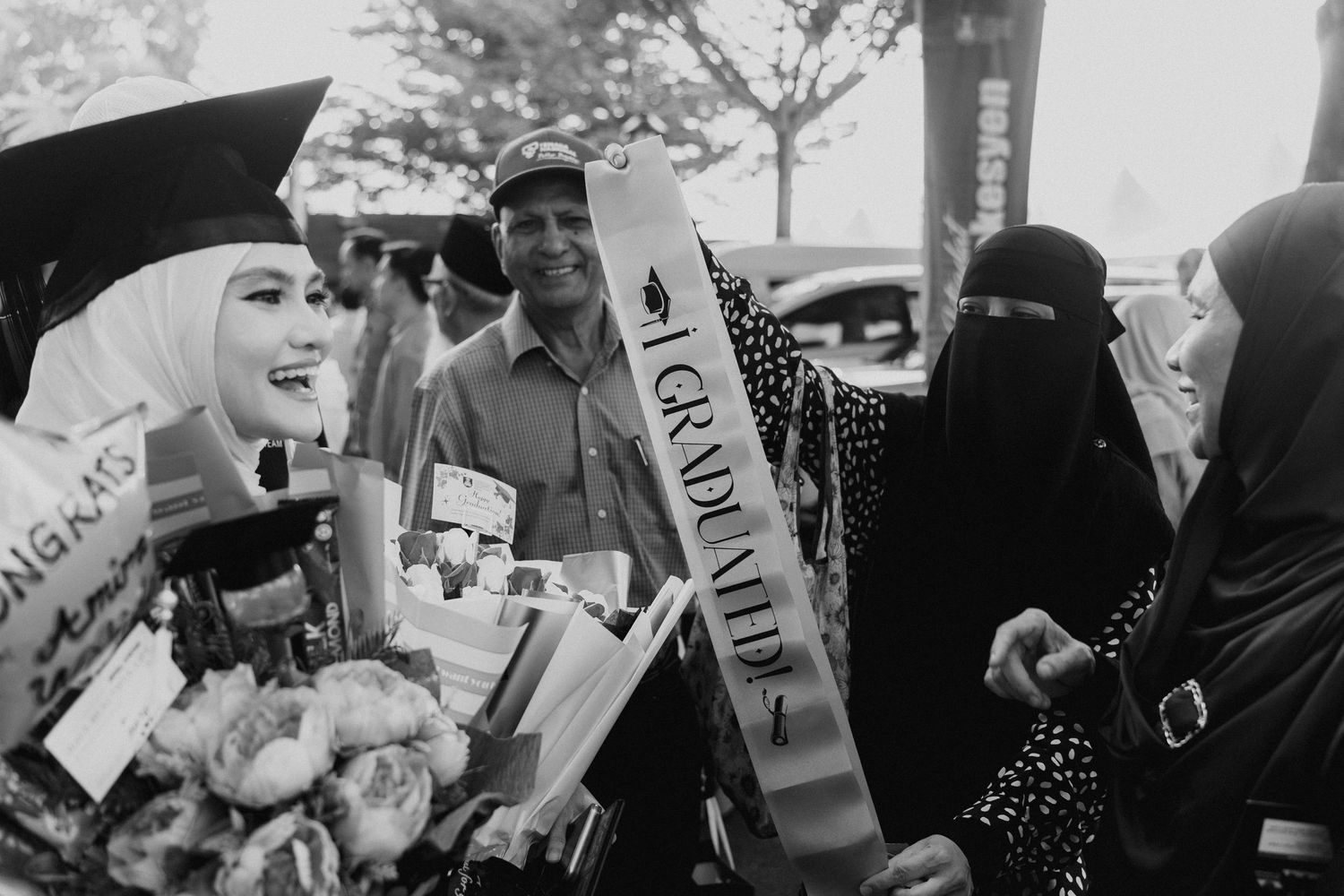 A graduate in graduation cap and gown stands with others wearing traditional religious garments at an outdoor ceremony.