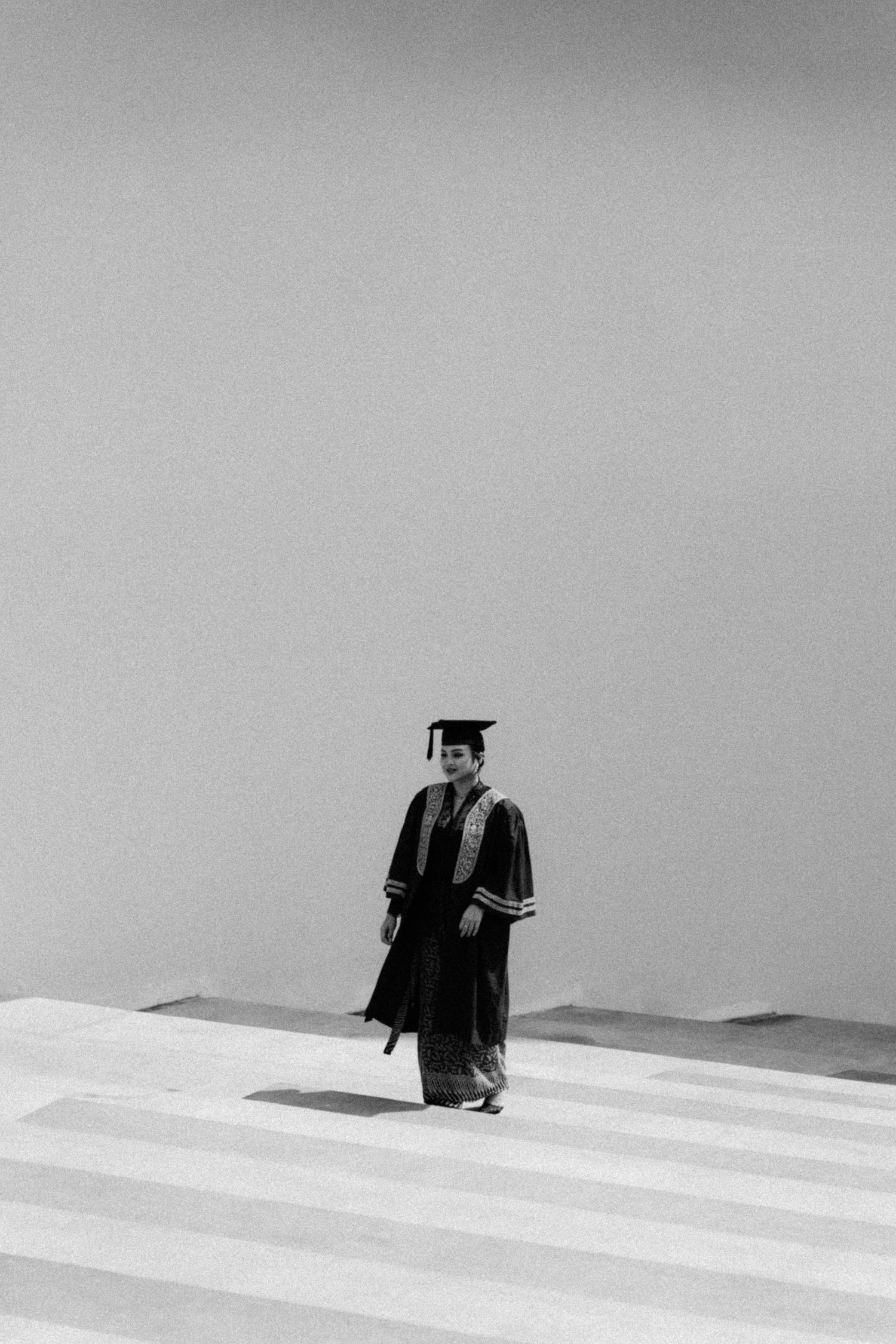 A graduate in traditional cap and gown stands atop a striped staircase in black and white photography.