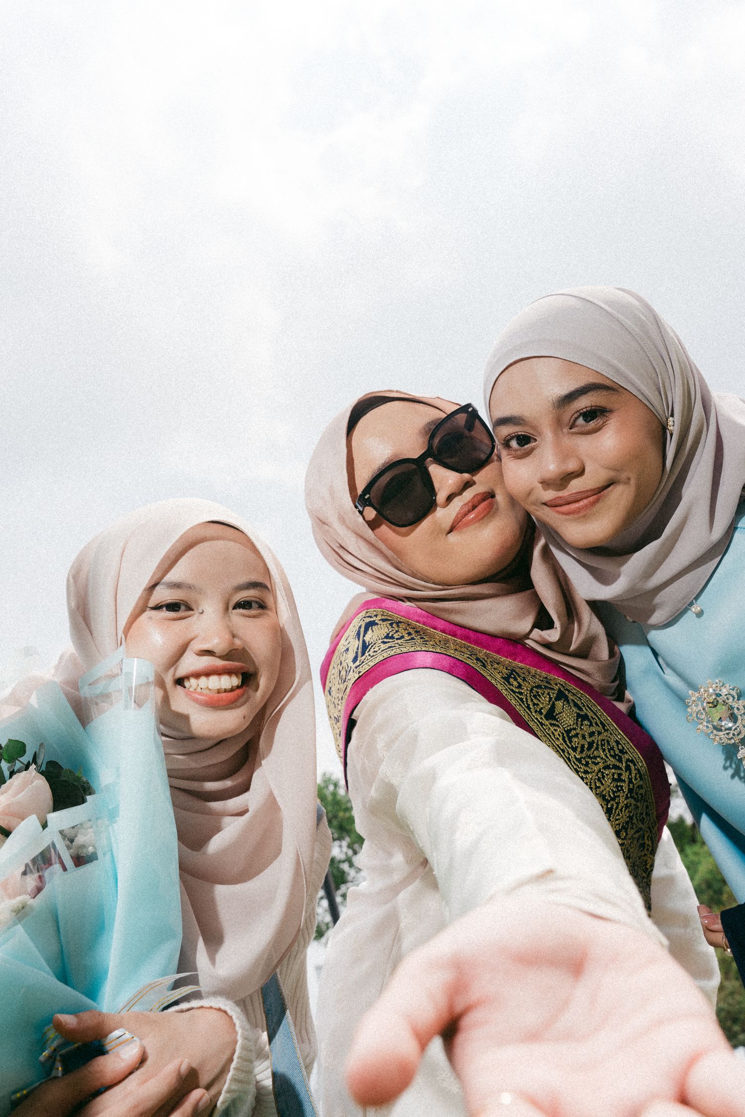 Three friends wearing hijabs and colorful clothing smile while taking a selfie together outdoors.