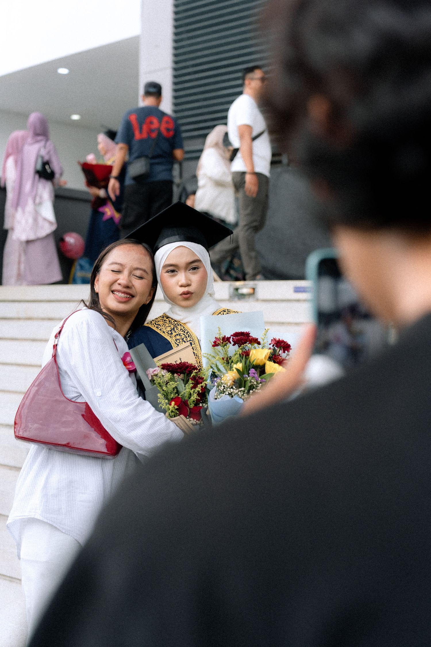 Students in white attire sit on steps with colorful flower bouquets during a graduation ceremony.