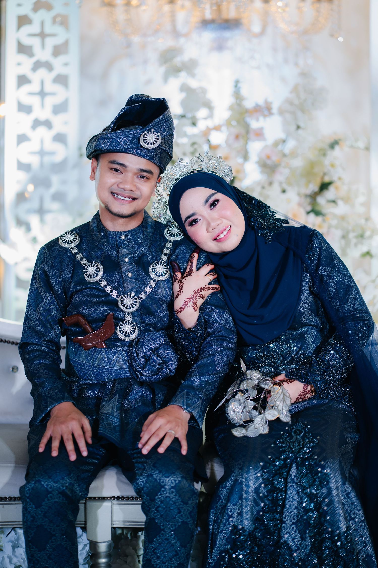 A couple in traditional Malaysian formal wear with matching navy blue and silver songket patterns sitting together.