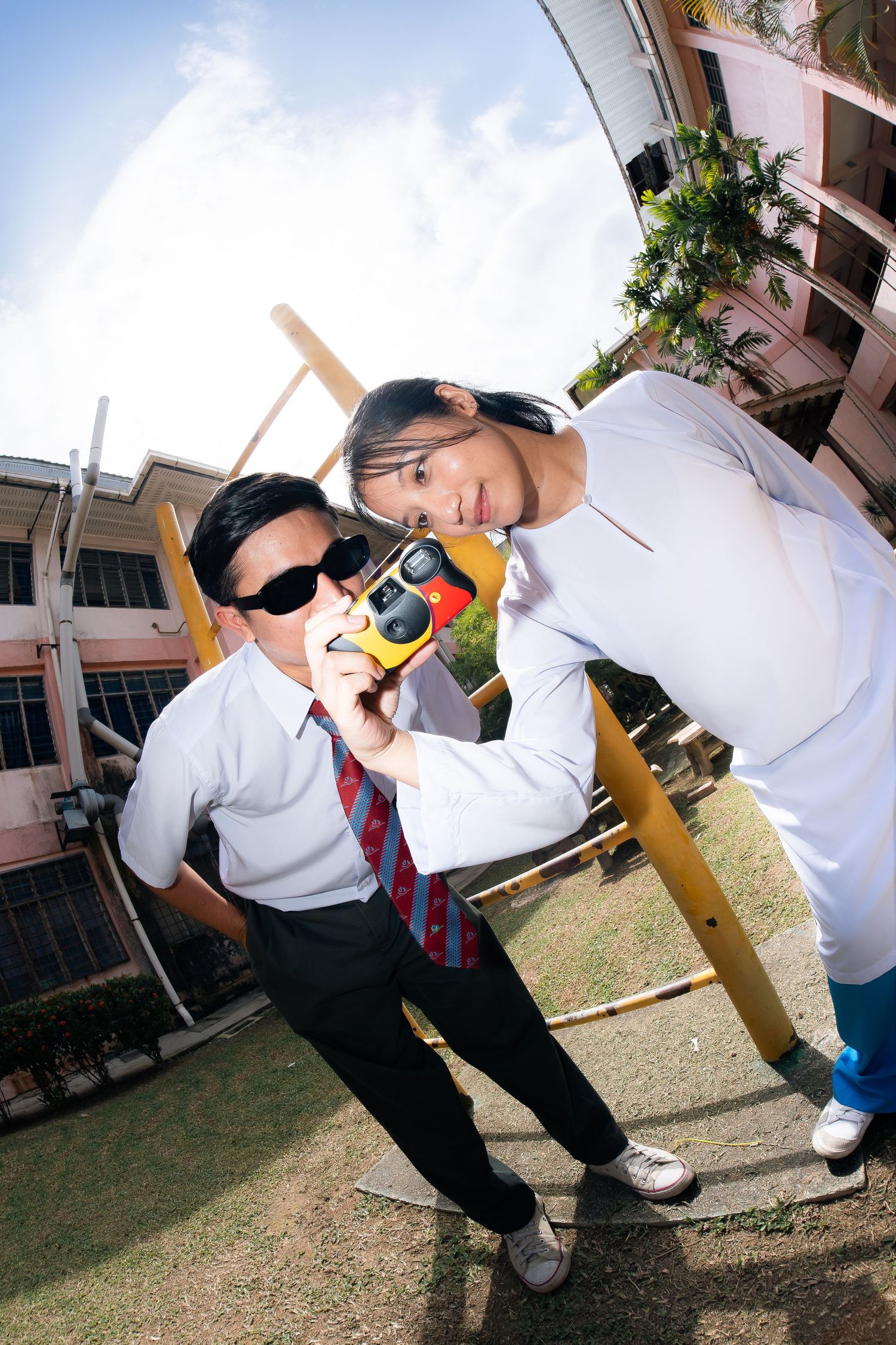 Two students in school uniforms pose playfully with baseball bats on a sunny day at their school campus.