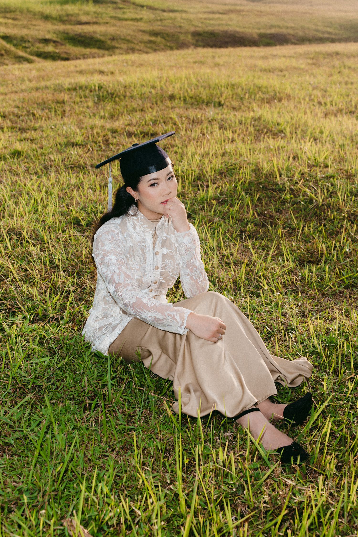 A person in a white lace top and khaki pants sits thoughtfully in a sunlit grassy meadow on a warm afternoon.