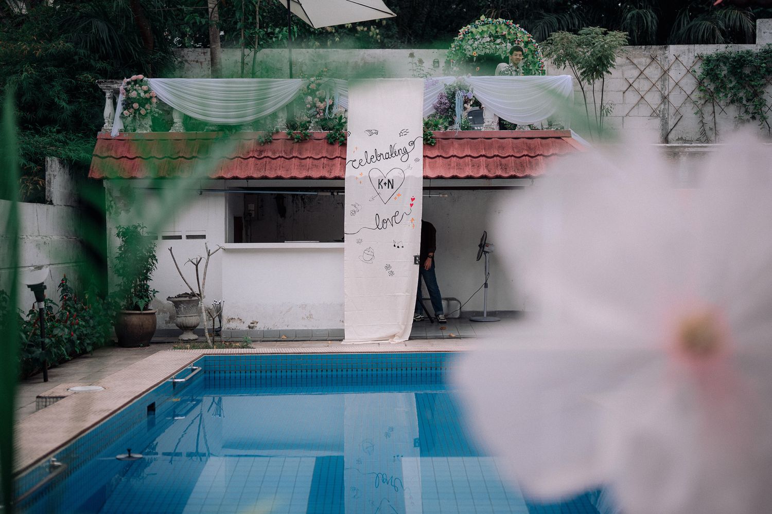 Outdoor swimming pool with a white banner sign, surrounded by tropical plants and a white flower in the foreground.