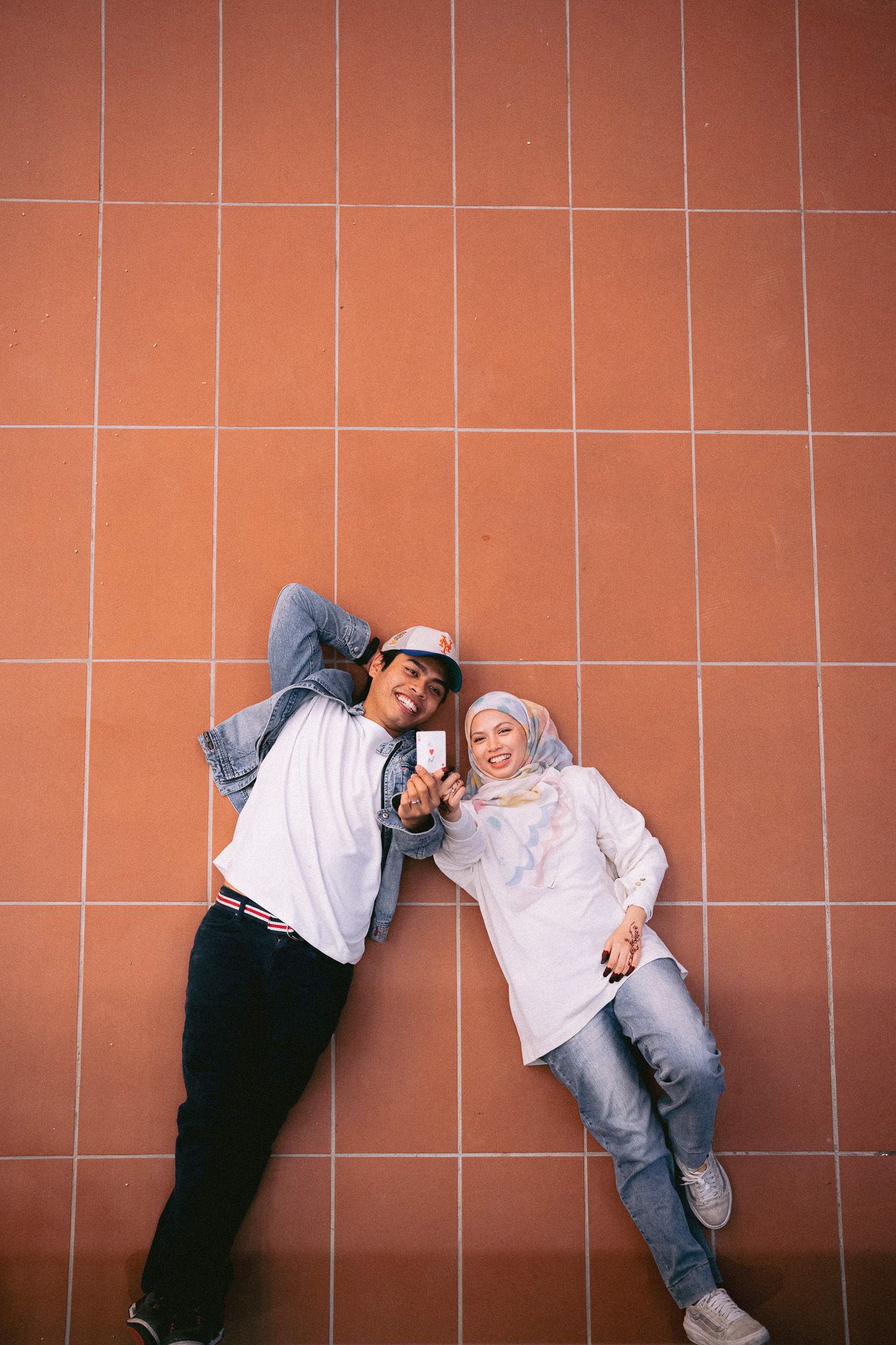 A couple leans against a terracotta tiled wall, smiling and taking a selfie, dressed in casual streetwear and hijab.