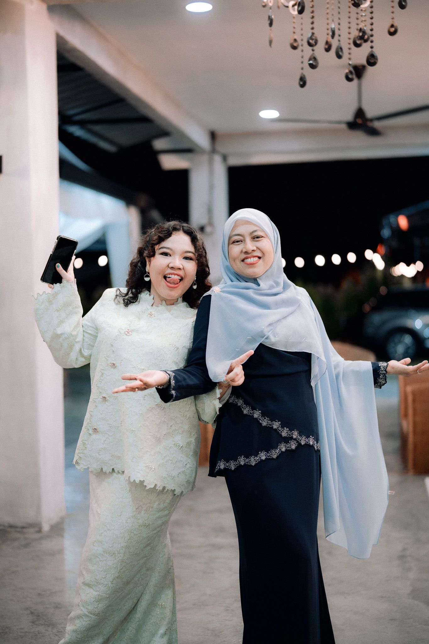 Two women in traditional Malay attire smiling and posing joyfully at an evening event.