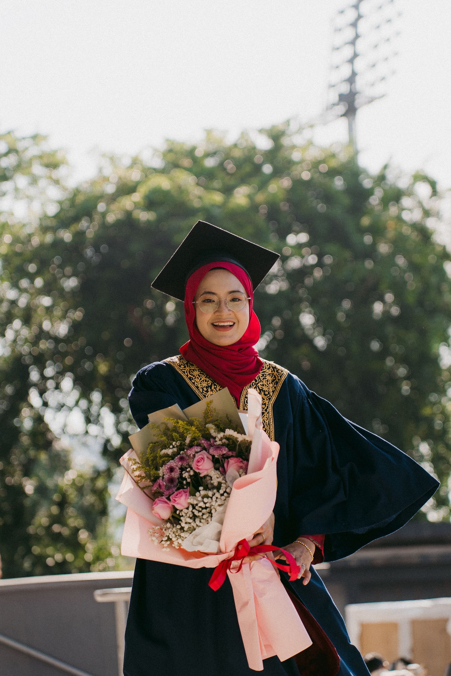 A graduate in academic regalia and red hijab holding a pink flower bouquet outdoors.