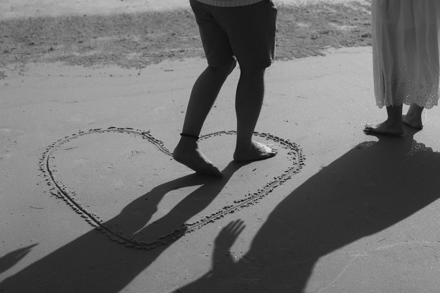 A heart shape drawn in sand with two people's shadows visible at sunset.