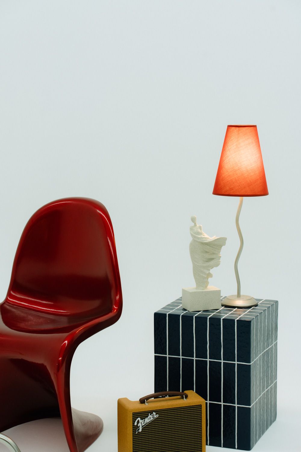 Red Panton chair beside tiled cube with white sculpture, red lamp, and Fender amp on light blue background.