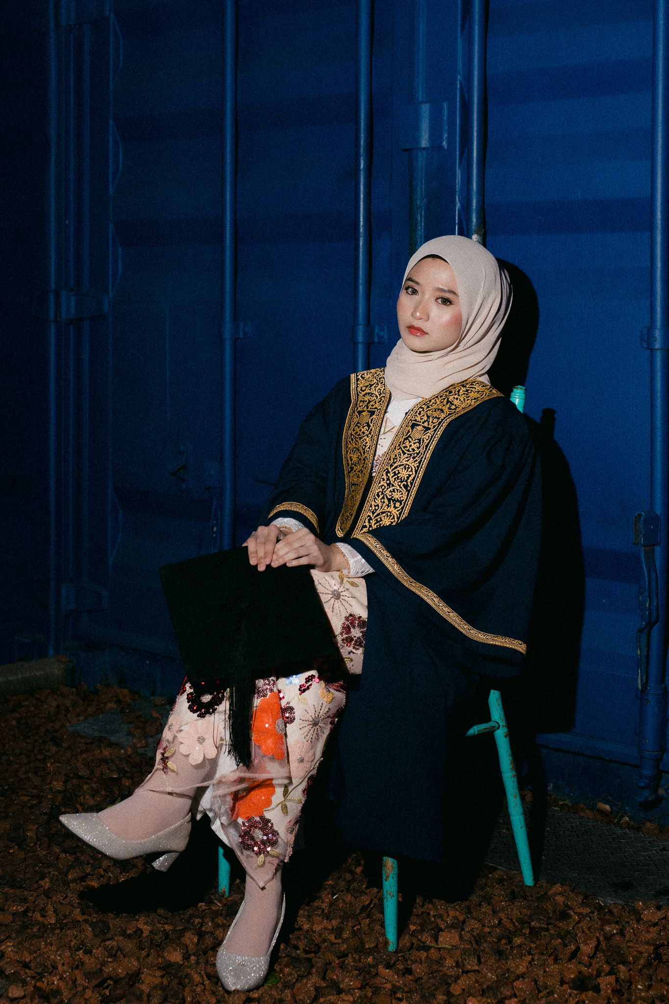 A woman in hijab and dark embroidered abaya sits gracefully on a chair against a blue wall.