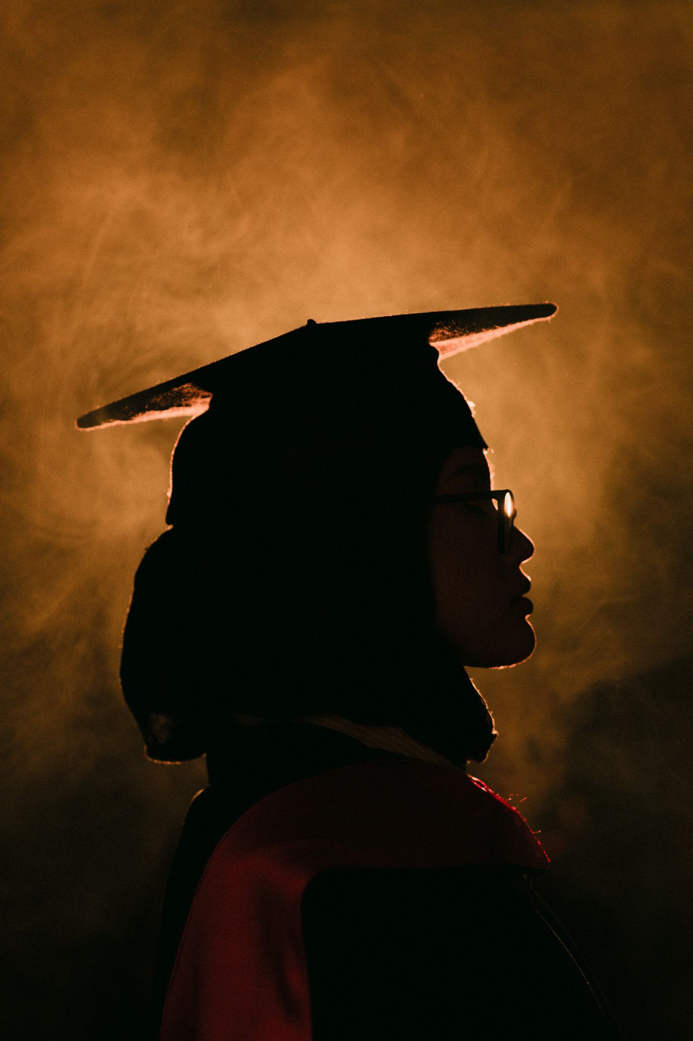 Silhouette of a graduate wearing mortarboard cap and gown against a dramatic golden smoky background.