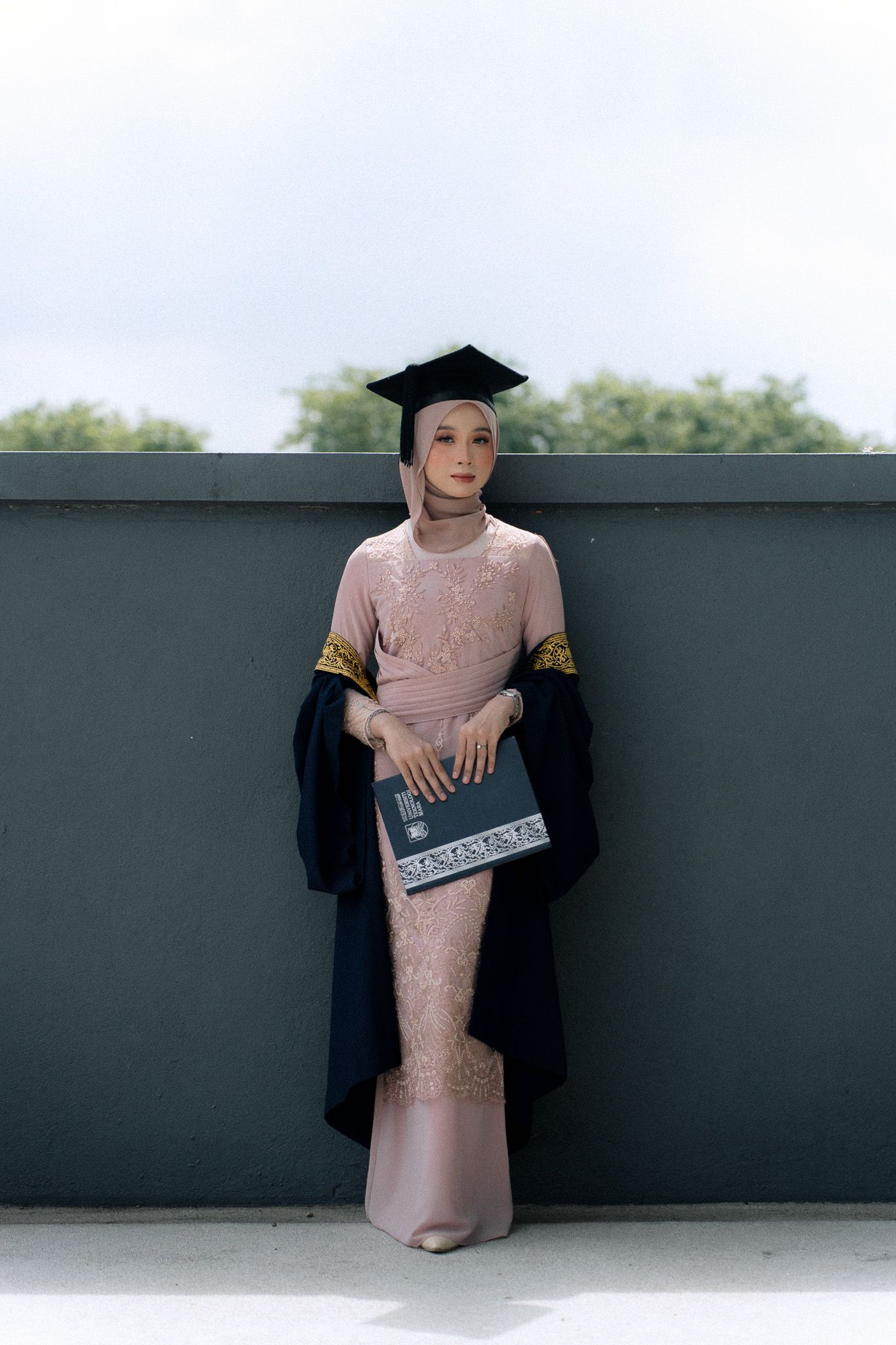 Graduate in pink dress and cap holding diploma standing against gray wall outdoors.