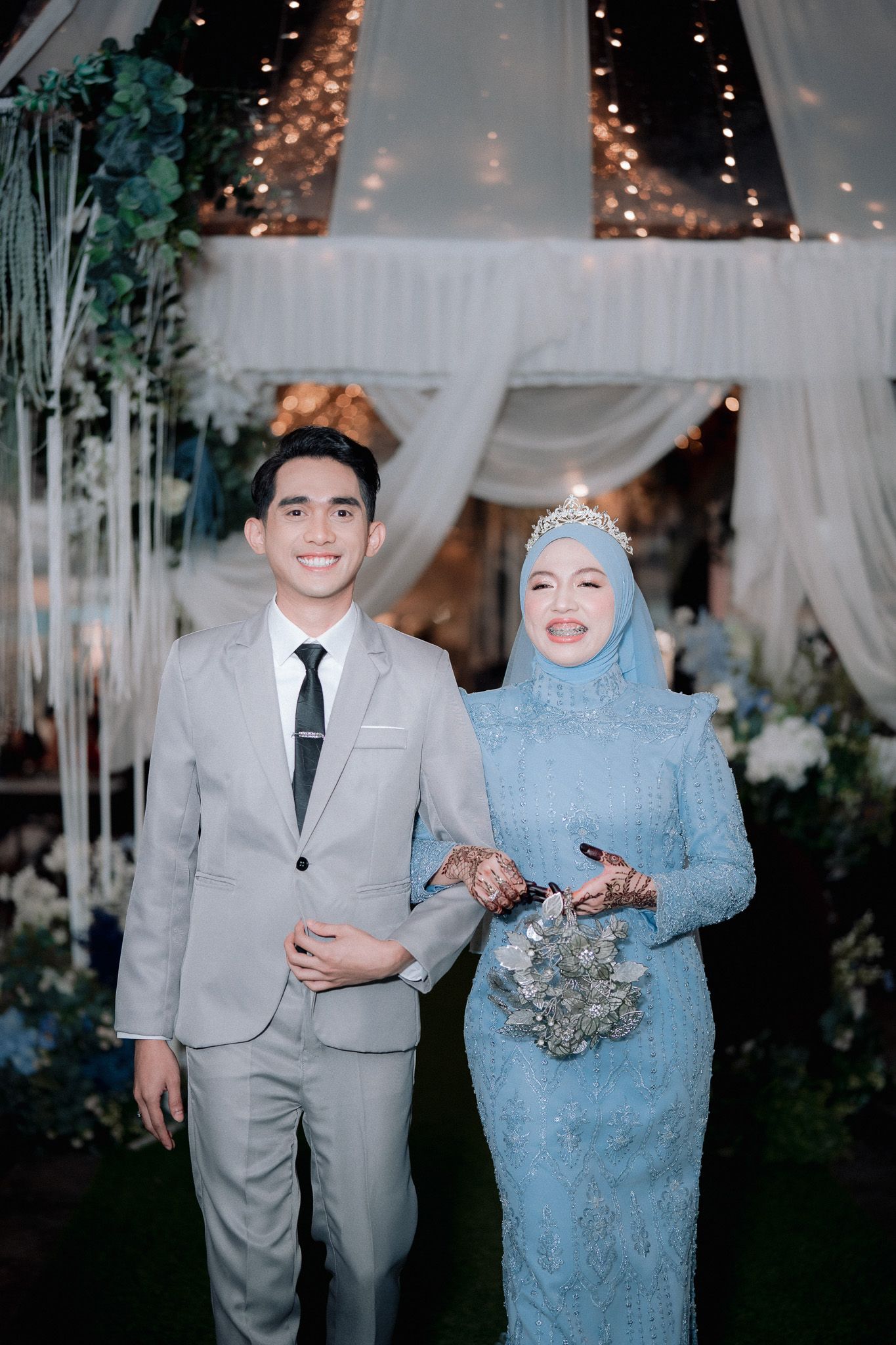 A couple in elegant wedding attire, groom in gray suit and bride in blue hijab gown with floral bouquet, posing at decorated venue.