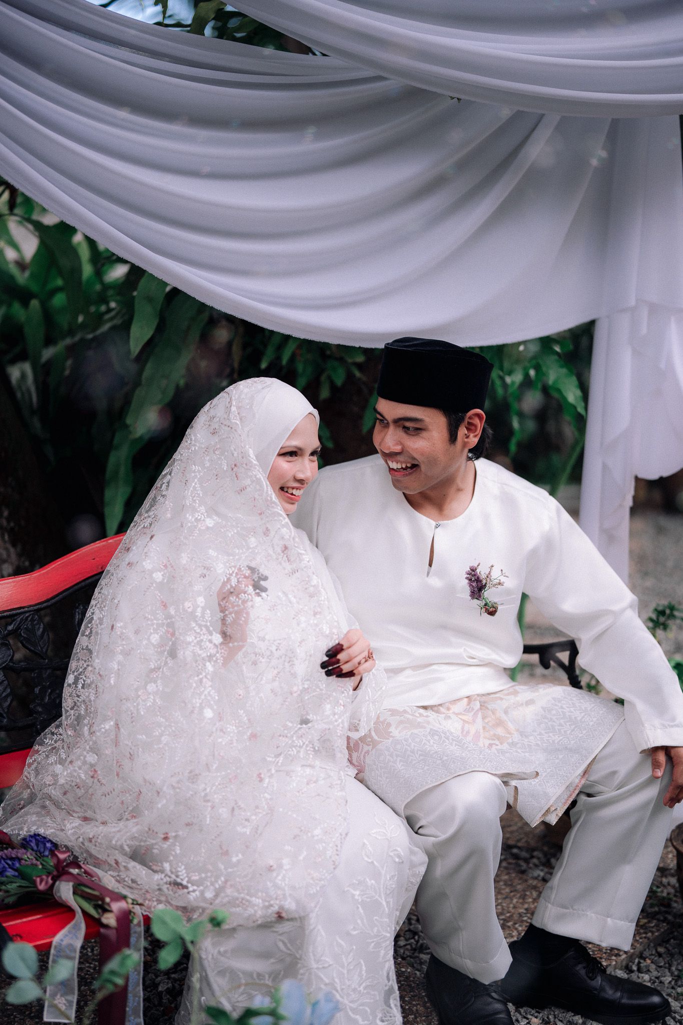 Muslim couple in traditional white wedding attire sitting together and smiling under a draped canopy outdoors.