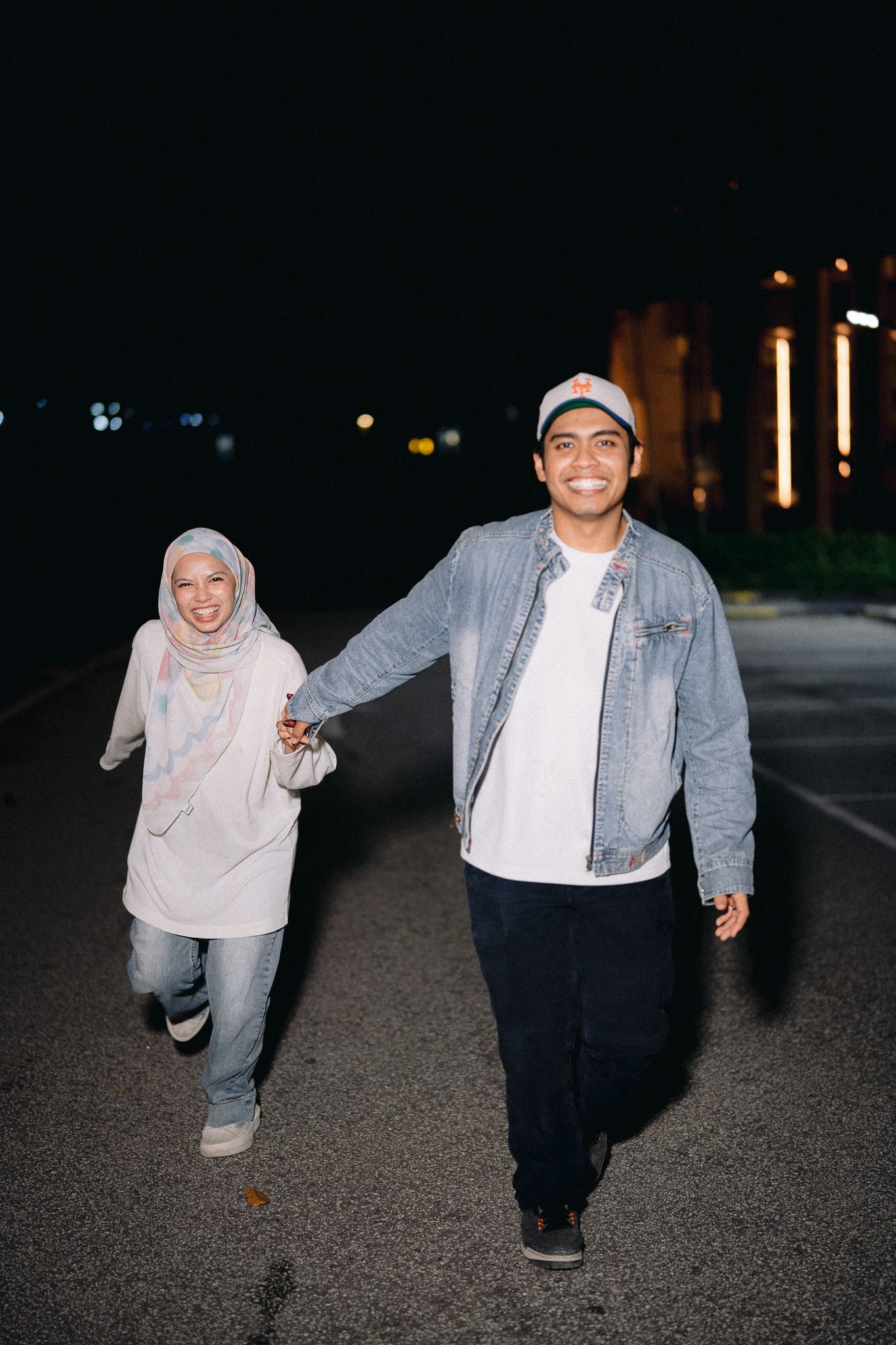 A smiling couple holds hands walking on a road at night, woman in hijab and man in denim jacket and cap.