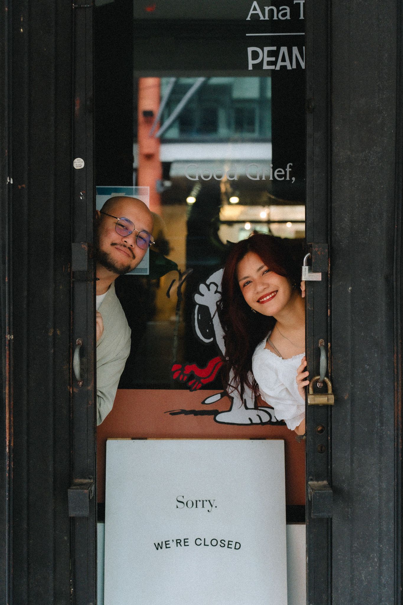 Two people smiling and peeking through a closed door with a Sorry We're Closed sign at a Peanuts-themed cafe.