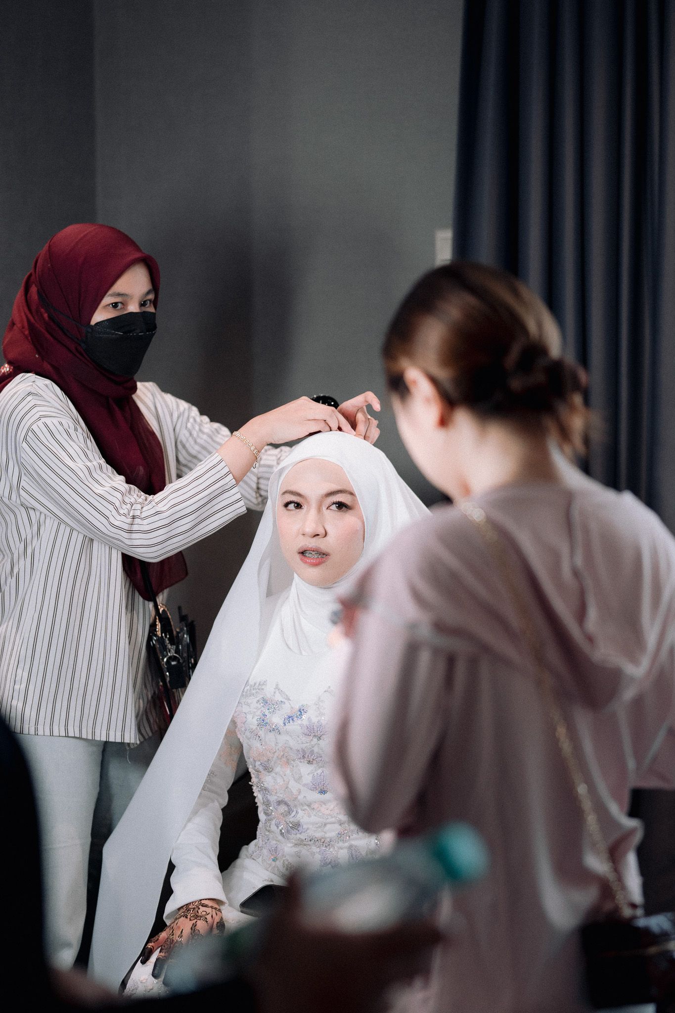 Muslim bride getting hijab styled by makeup artist in white wedding dress with henna hands.