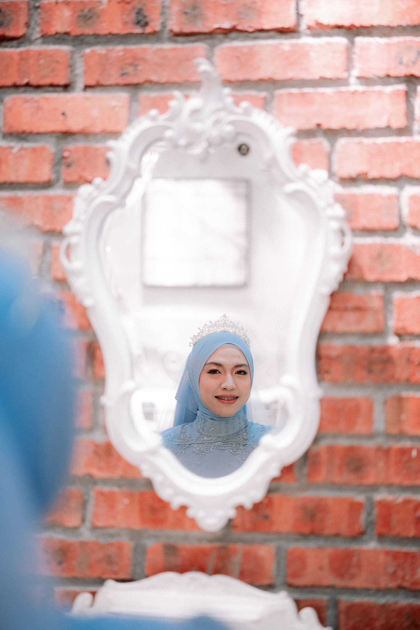 A woman in a blue hijab and crown reflected in an ornate white mirror against a red brick wall.
