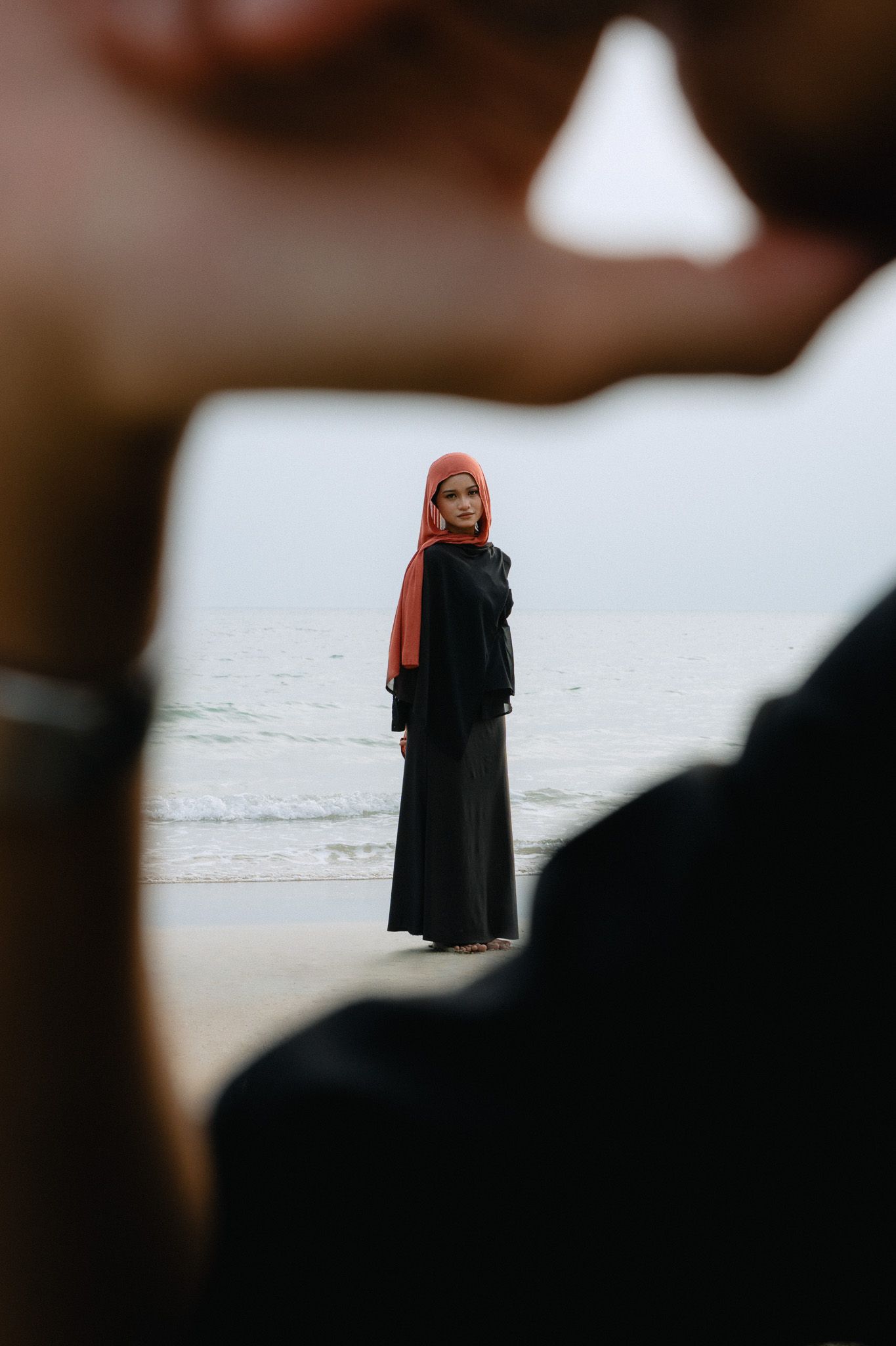 Woman in black abaya and red hijab standing on a beach, framed by blurred foreground figures.