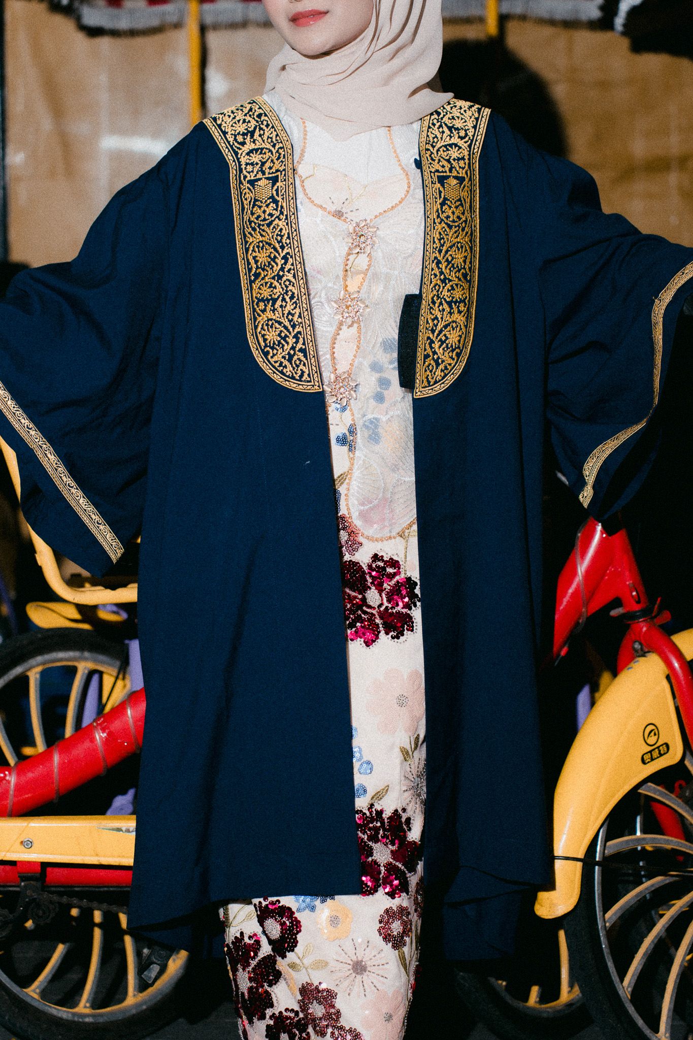 Woman wearing navy blue abaya with gold embroidery over floral dress and cream hijab, standing outdoors.
