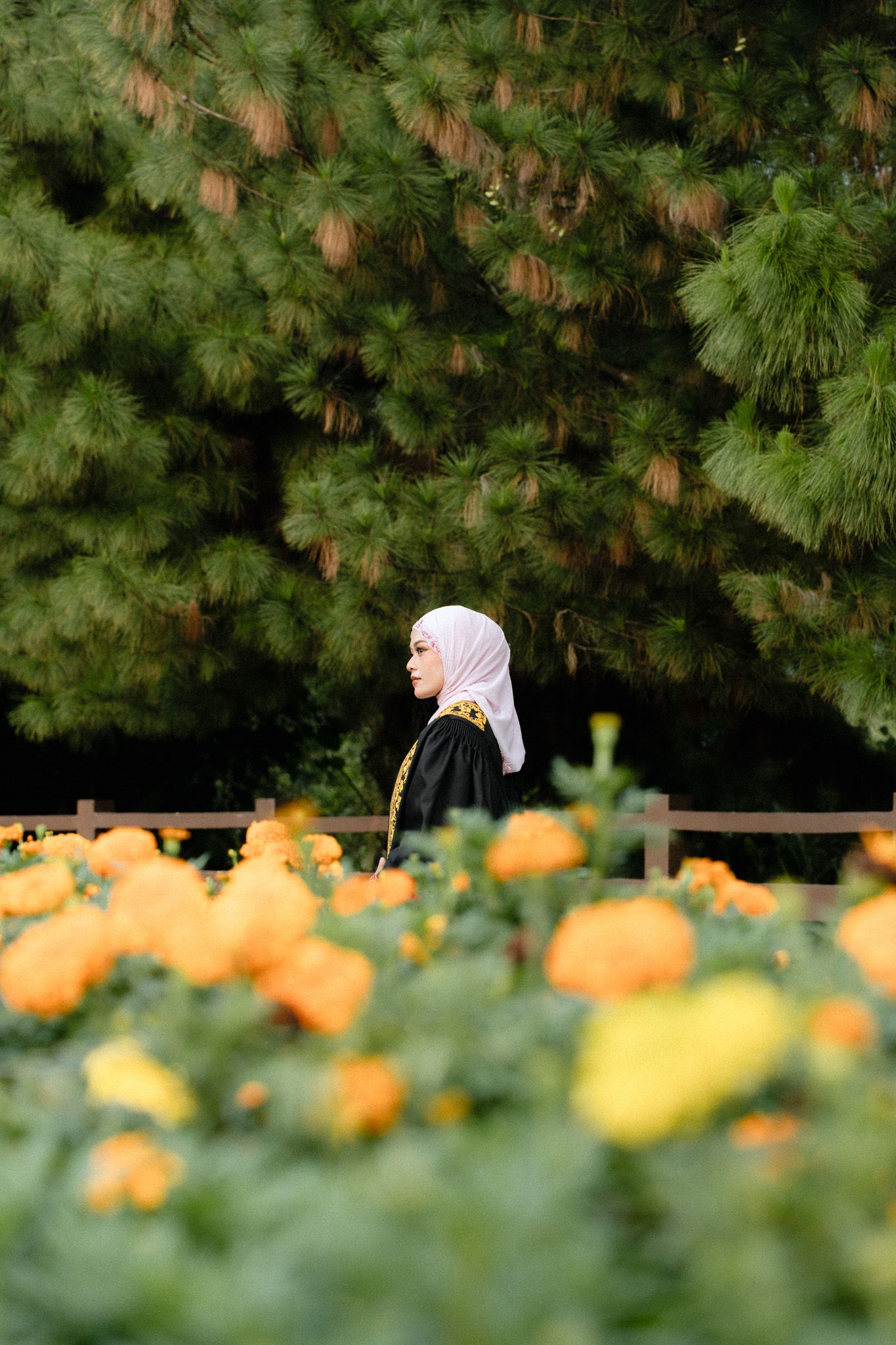 Woman in hijab and black graduation gown standing among orange and yellow flowers with green pine trees behind.