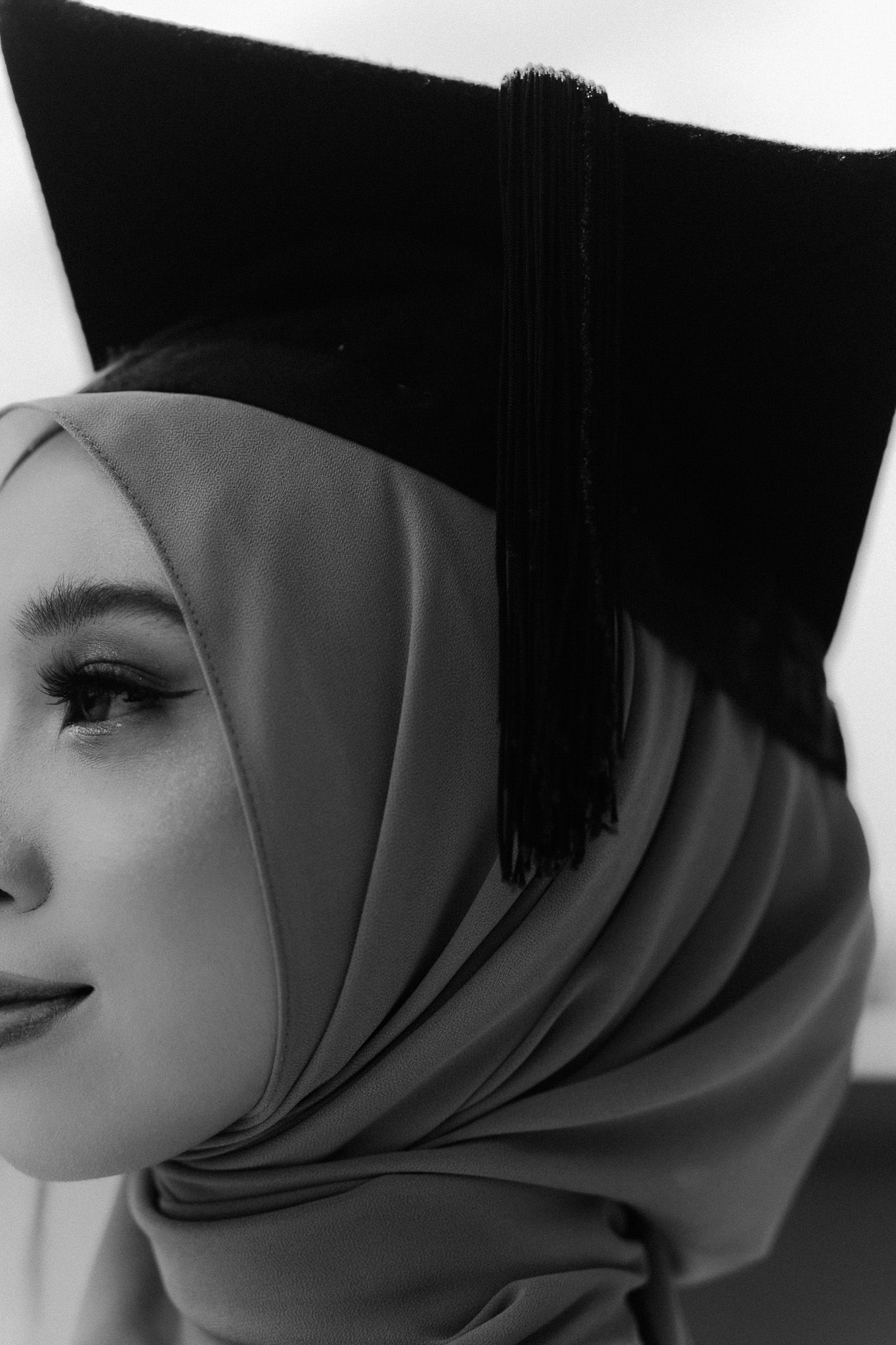 Black and white portrait of a young woman wearing a hijab and graduation cap with tassel, celebrating academic achievement.