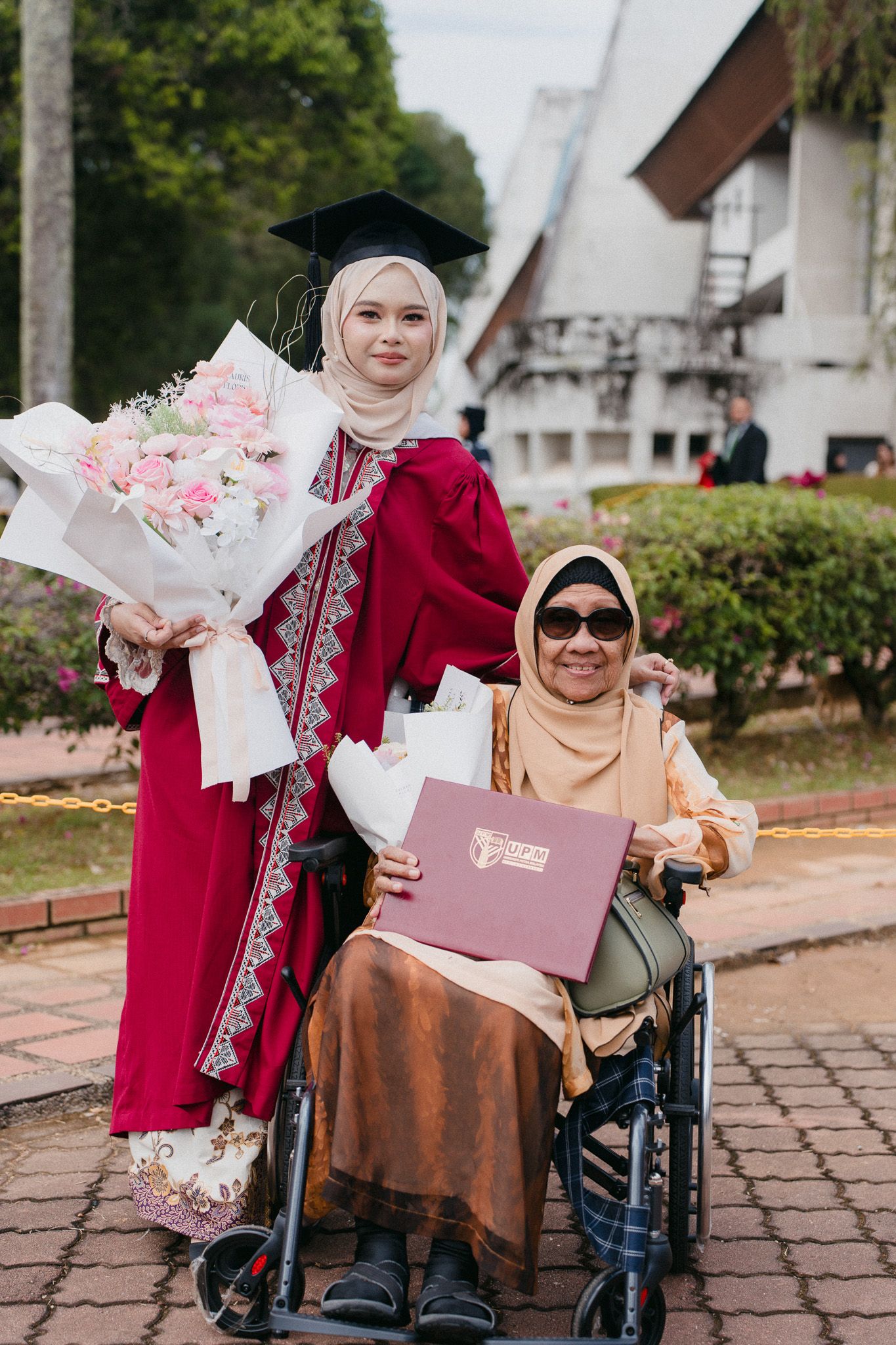 Muslim graduate in red robe holding flowers poses with elderly woman in wheelchair holding diploma outdoors.