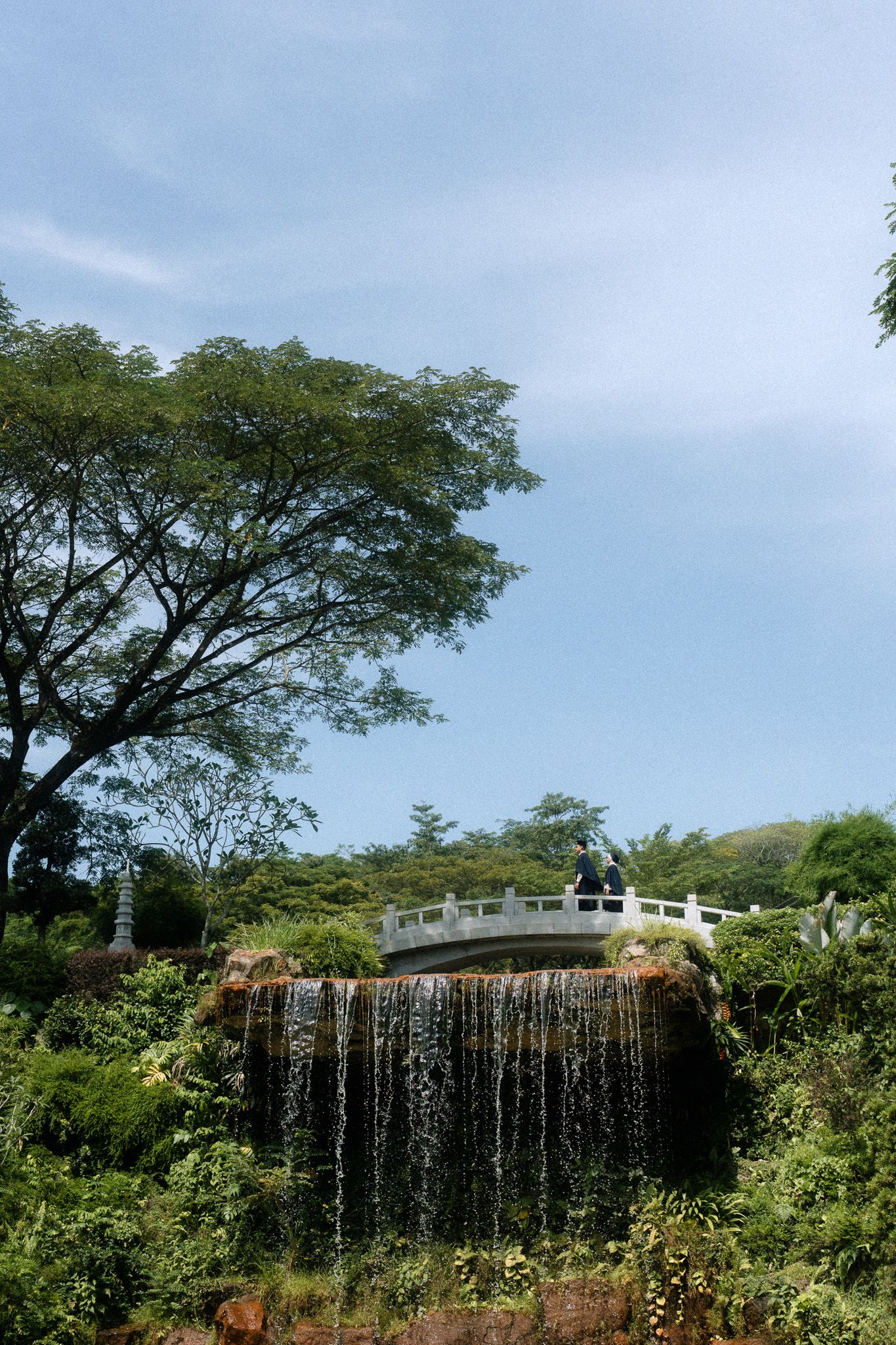 A serene garden waterfall cascades over rocks beneath an ornate white bridge, surrounded by lush greenery and a clear blue sky.