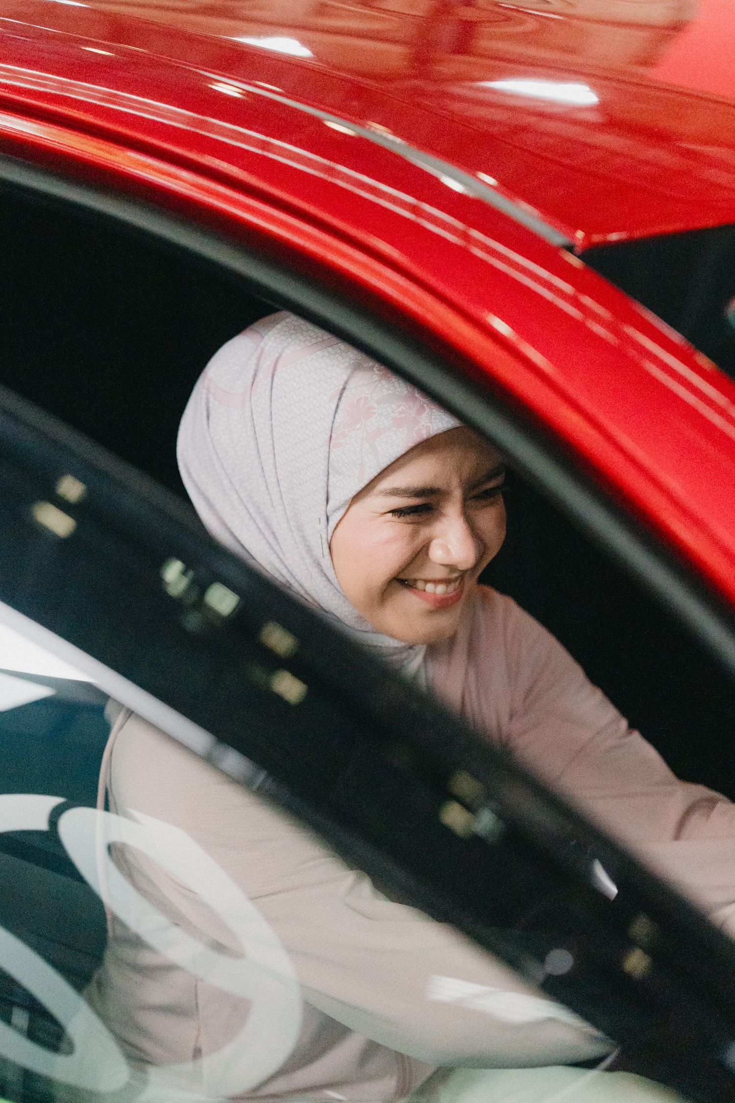A woman in a hijab sits in a bright red car, smiling through the window frame.