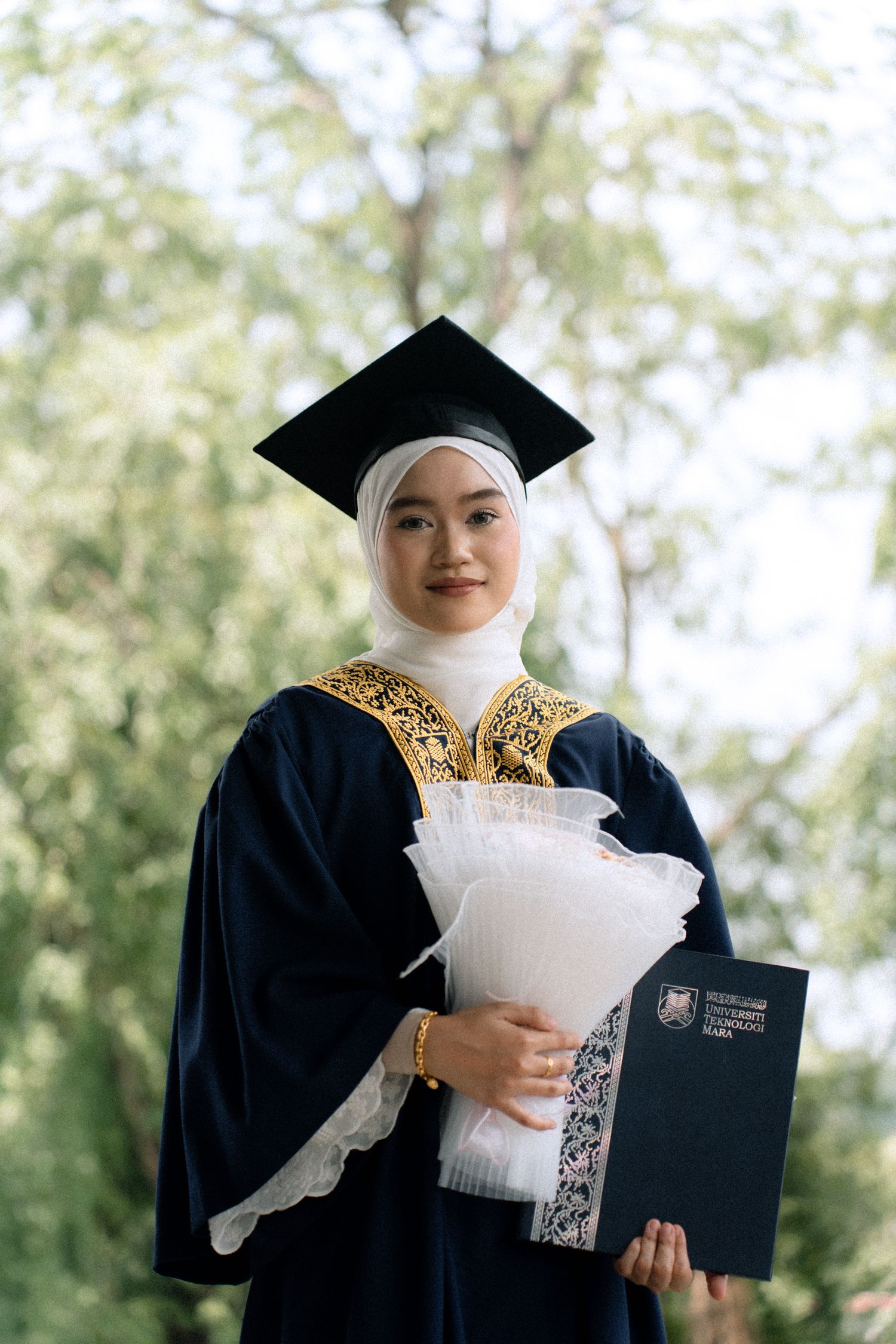 Graduate in black cap and gown holding diploma and white flowers during outdoor graduation ceremony.