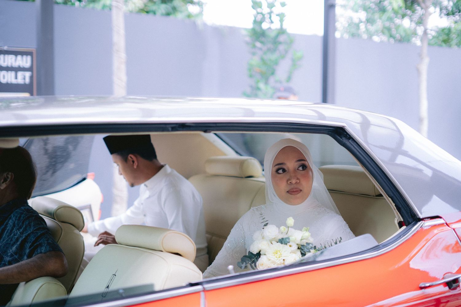 Someone in white sits in the back seat of an orange classic car holding a white bouquet.