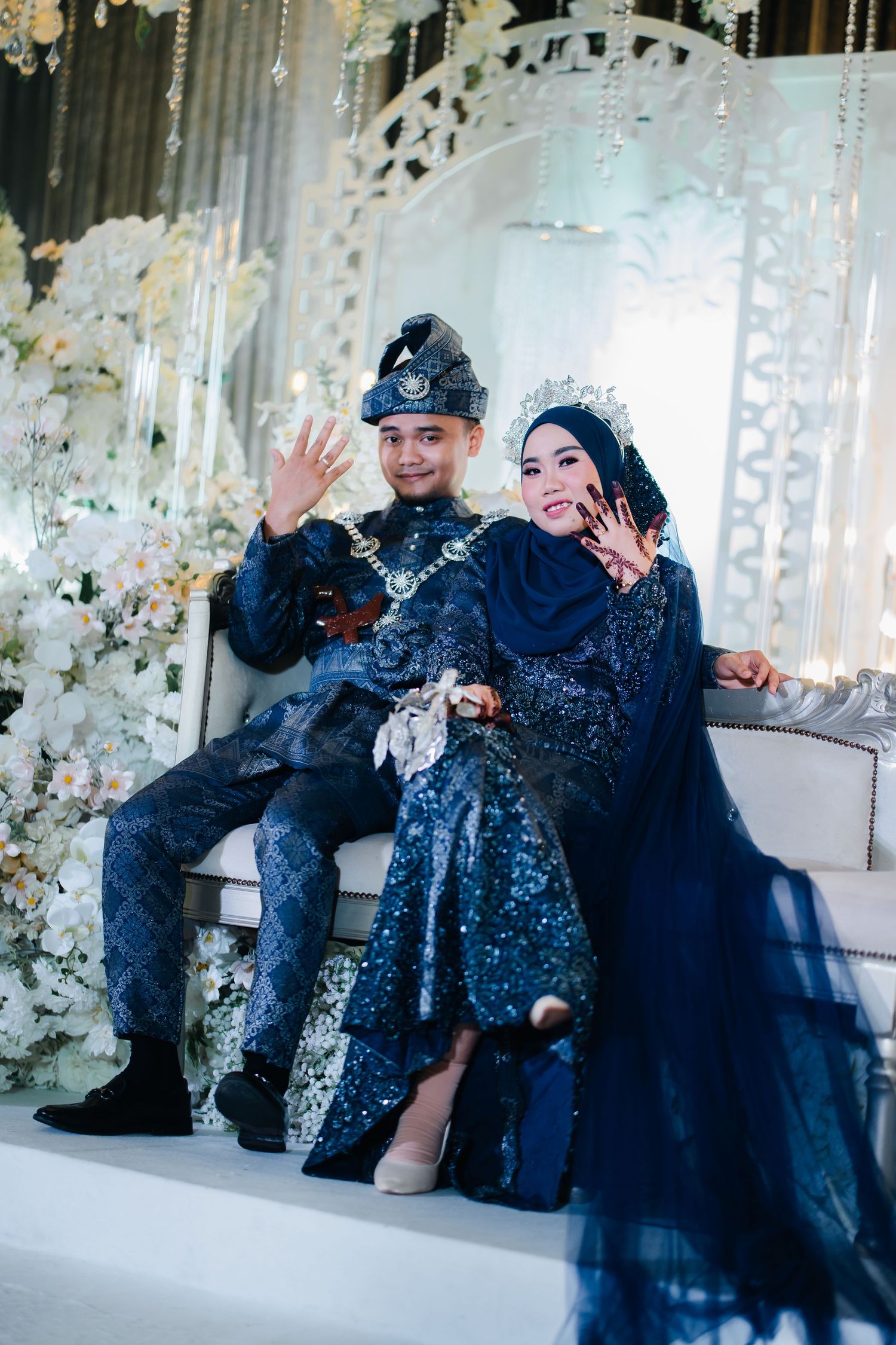 Two people in elegant blue and black formal attire pose playfully on a white couch with floral decorations in the background.