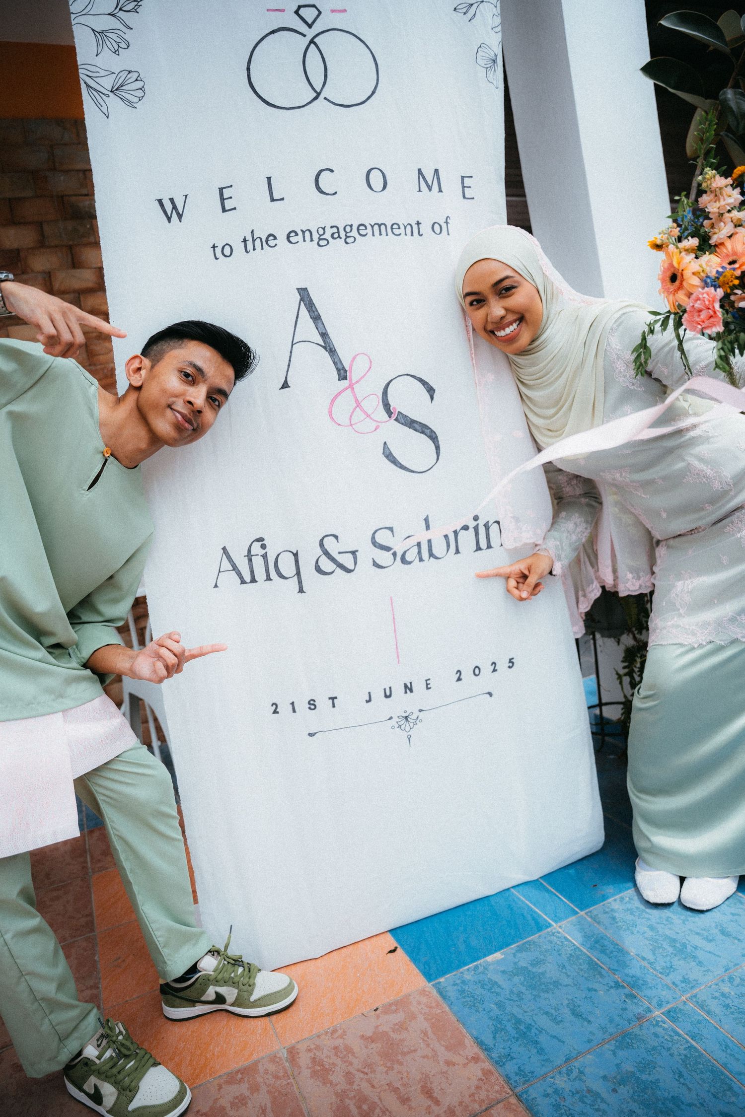 Two people in pastels pose beside a white engagement welcome sign with interlocked rings design on blue carpet.