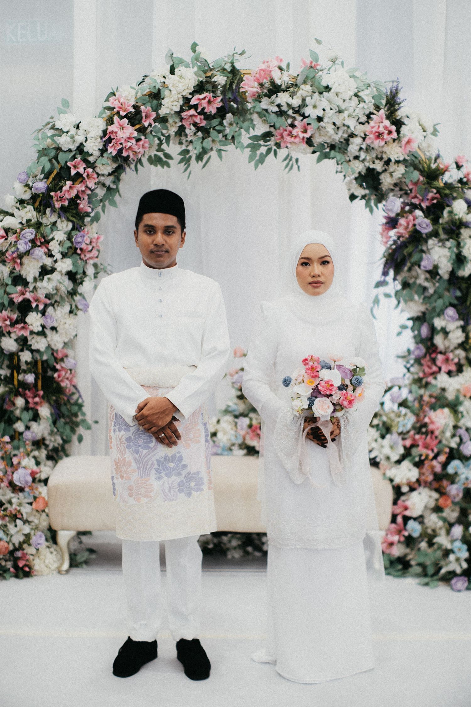 Elegant Malaysian wedding photography with floral arch backdrop featuring soft pink roses and white flowers during ceremony.