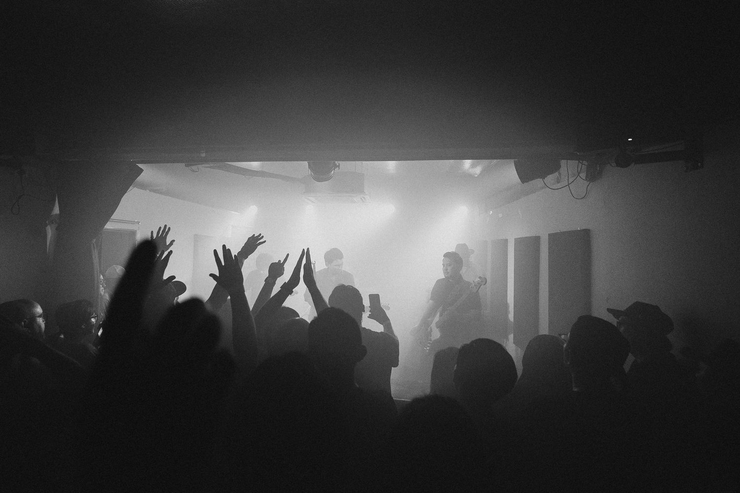 A silhouetted crowd with raised hands enjoys a concert in a dark venue with dramatic fog and lighting effects.