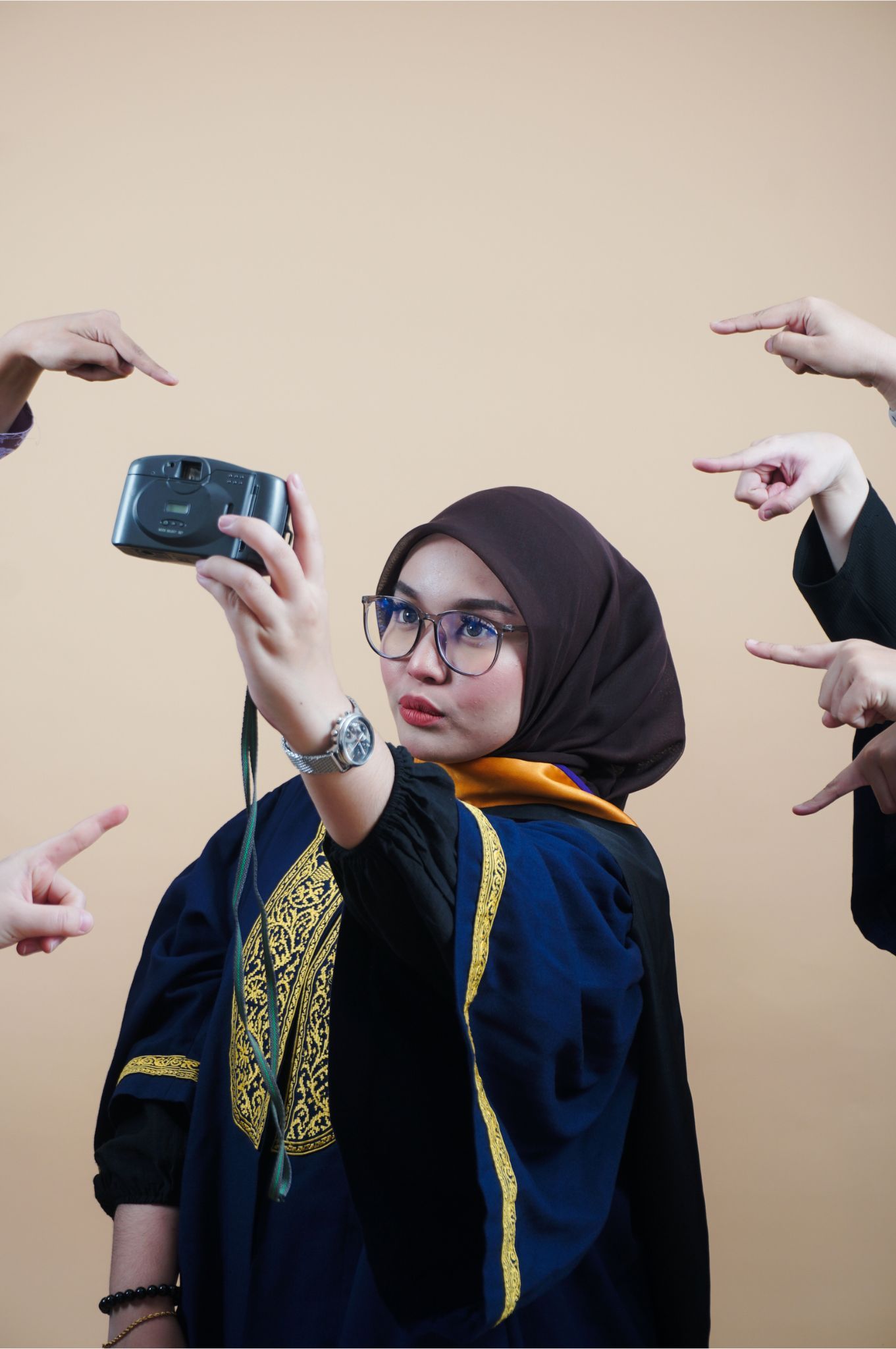 A young woman in graduation robes and hijab takes a selfie with a camera while multiple hands point at her.