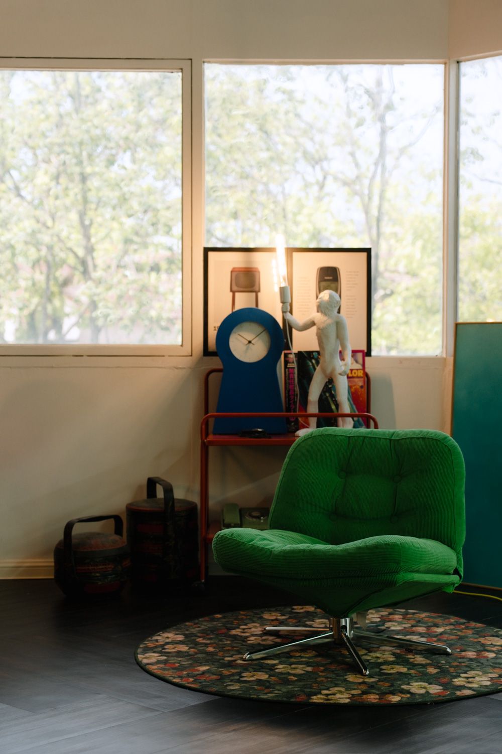 Retro green velvet swivel chair on floral rug beside a shelf with blue clock and white figurine near large windows.