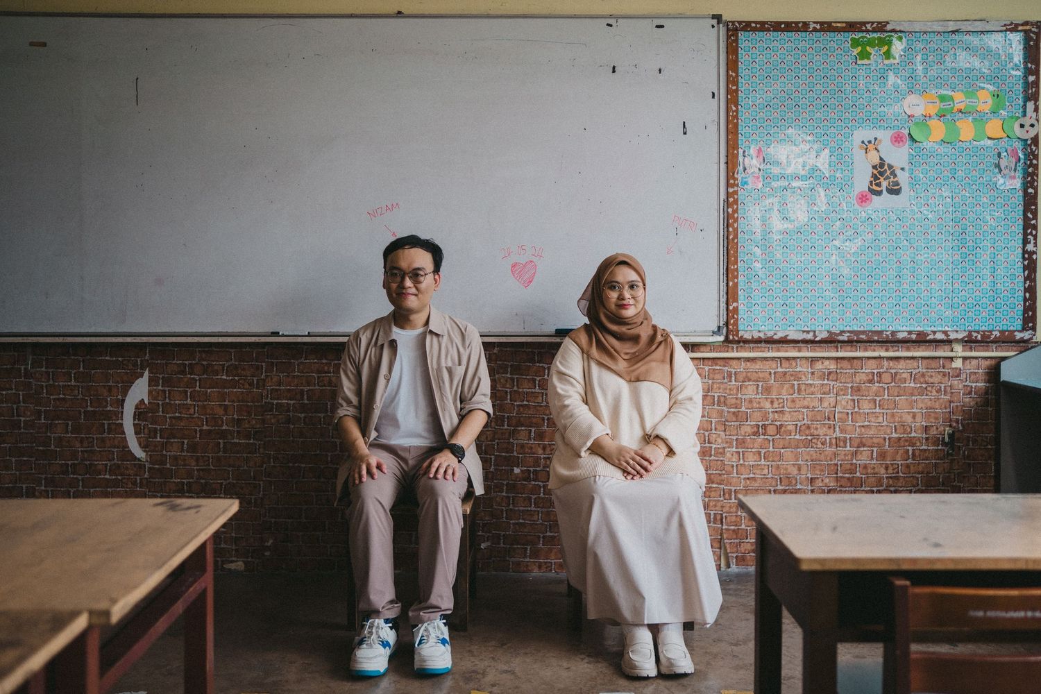 Two students posing against brick wall in classroom with whiteboard and bulletin board visible.