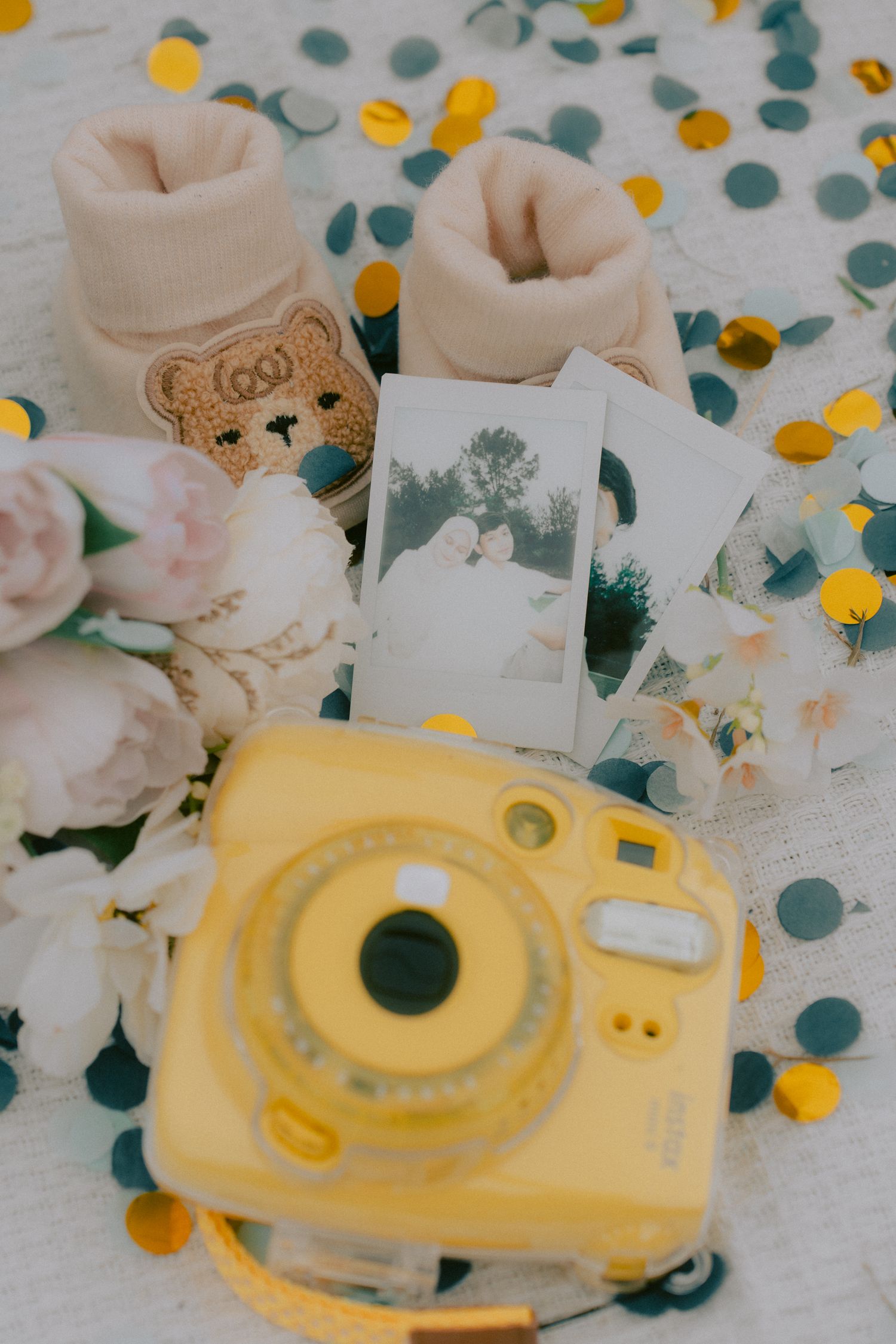 Yellow instant camera, teddy bear booties, and polaroid photos arranged with confetti on white fabric.