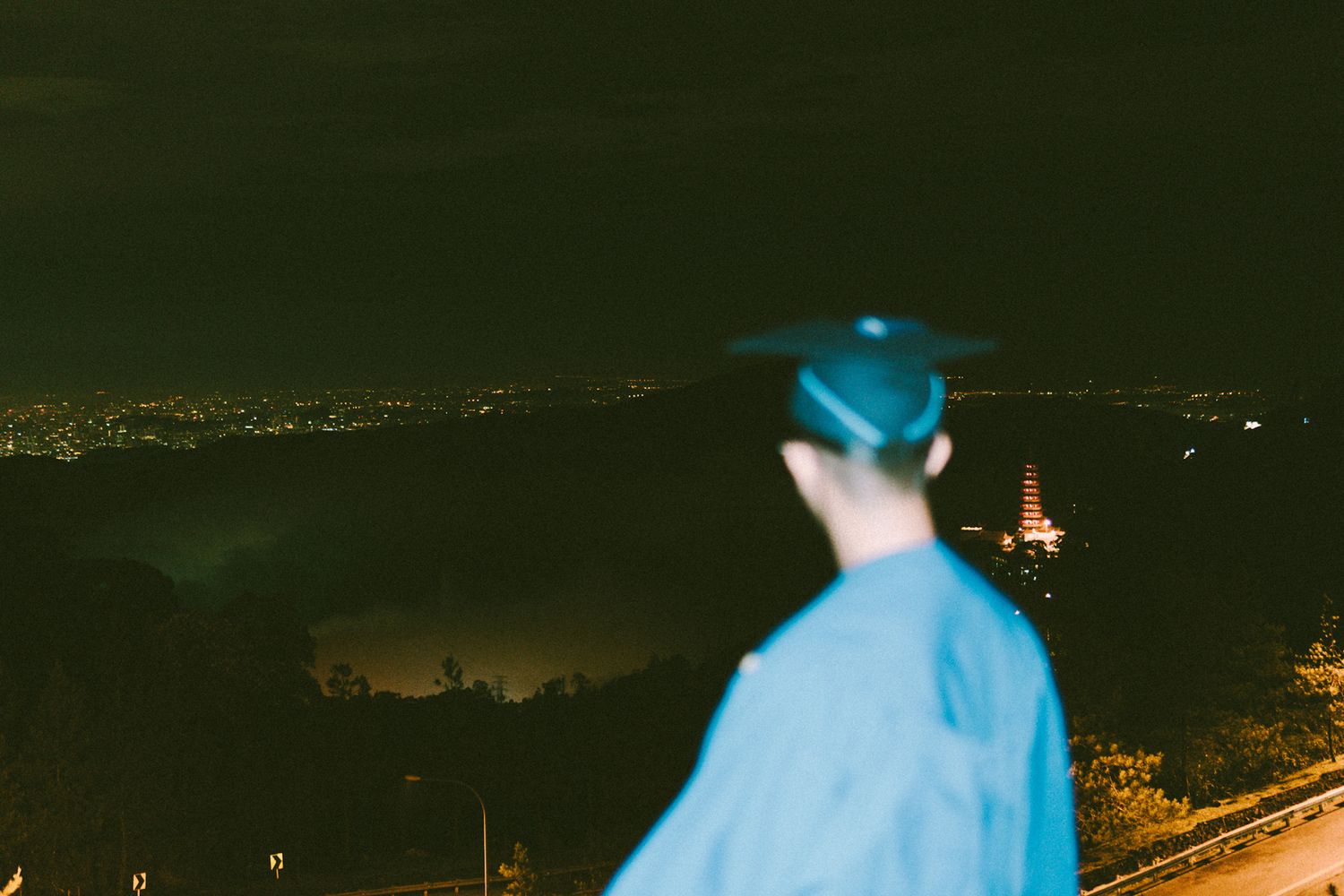 Blurred figure in blue overlooking city lights and distant scenery at night.
