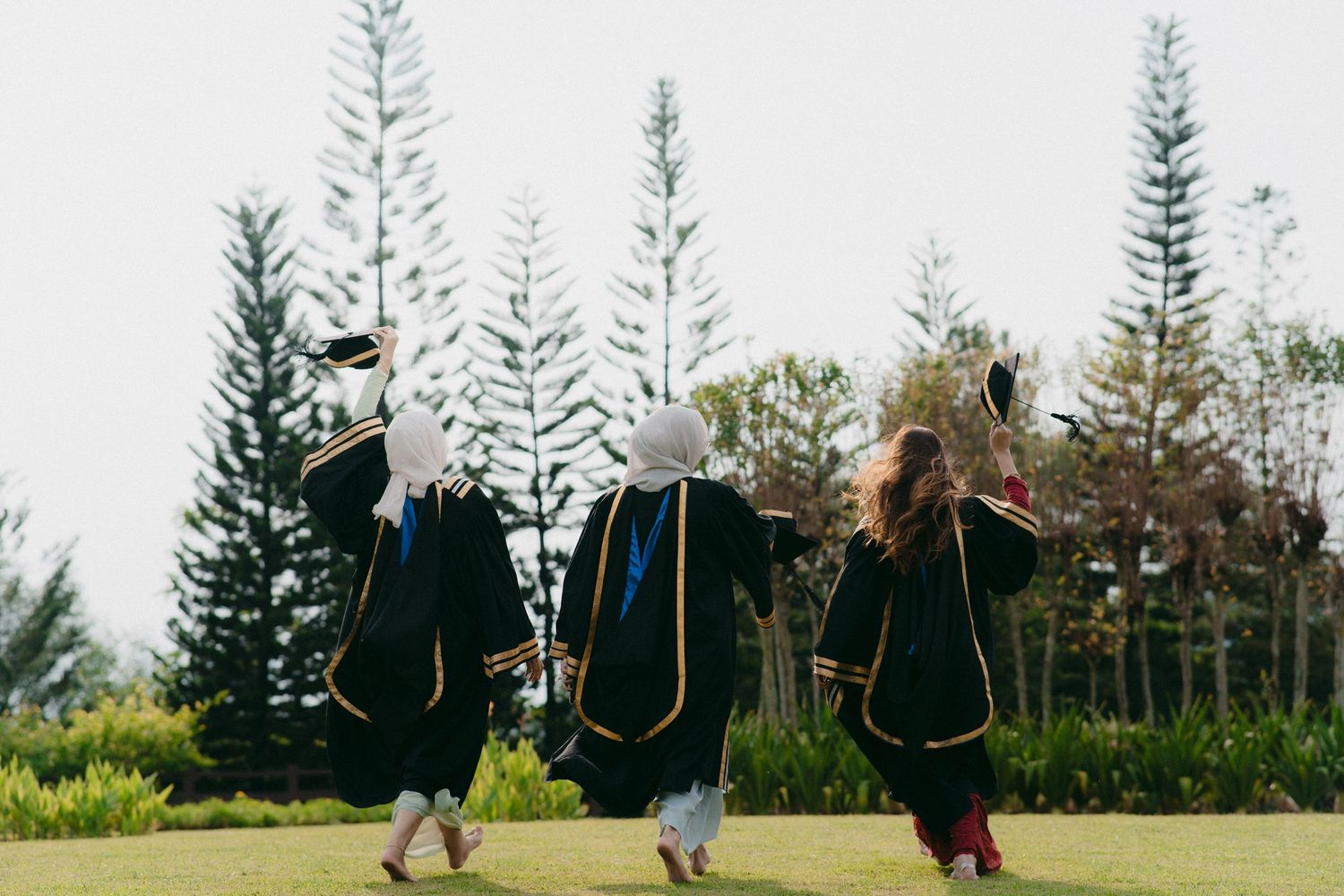 Three graduates in black and gold academic regalia dance together on a lawn with pine trees.