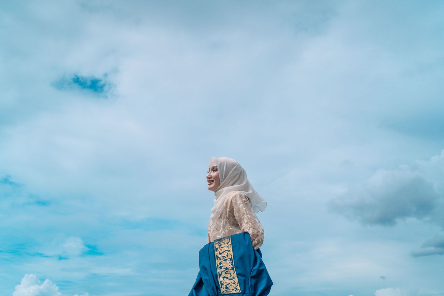 Graduate in hijab poses against cloudy blue sky background.