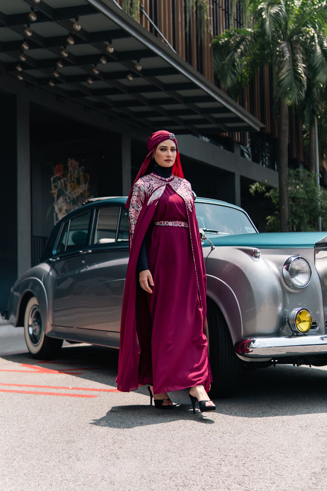 Burgundy modest dress and hijab styled against vintage car in modern architectural setting.