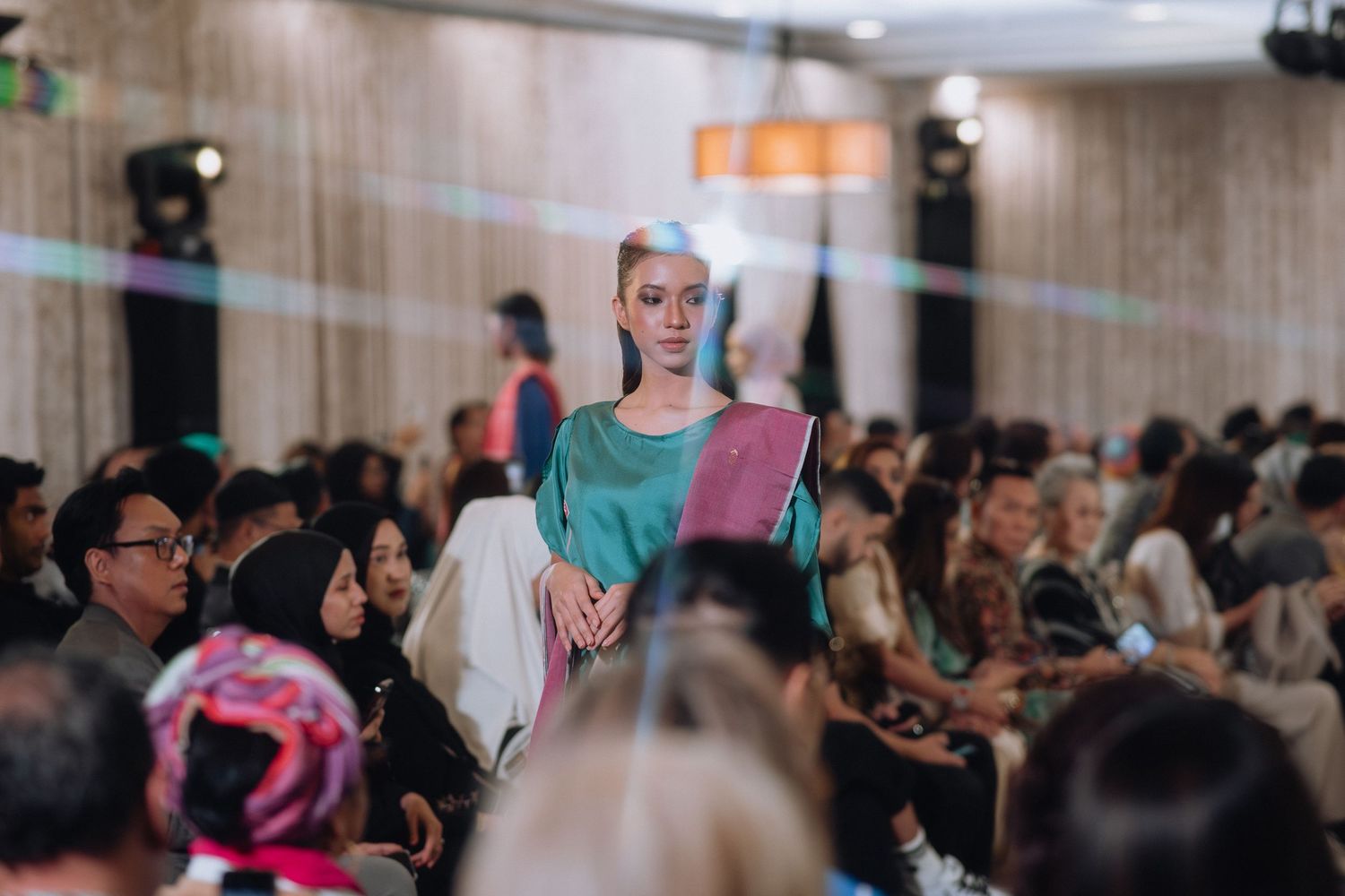 Model presents teal and white fashion design on runway at indoor fashion show.