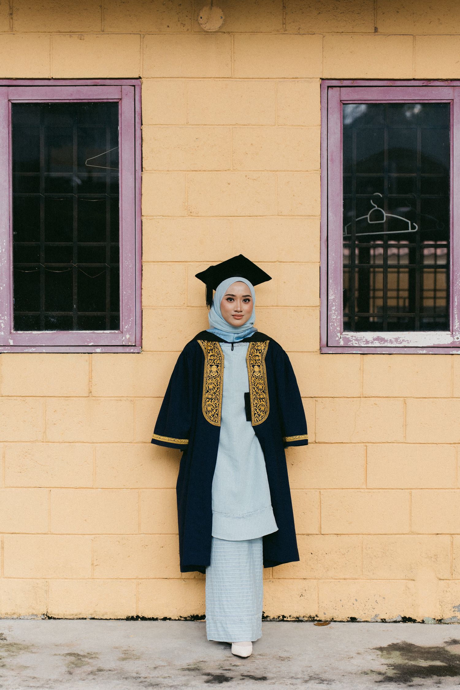 Graduate wearing traditional black and gold academic regalia with light blue attire poses against yellow wall with purple windows.